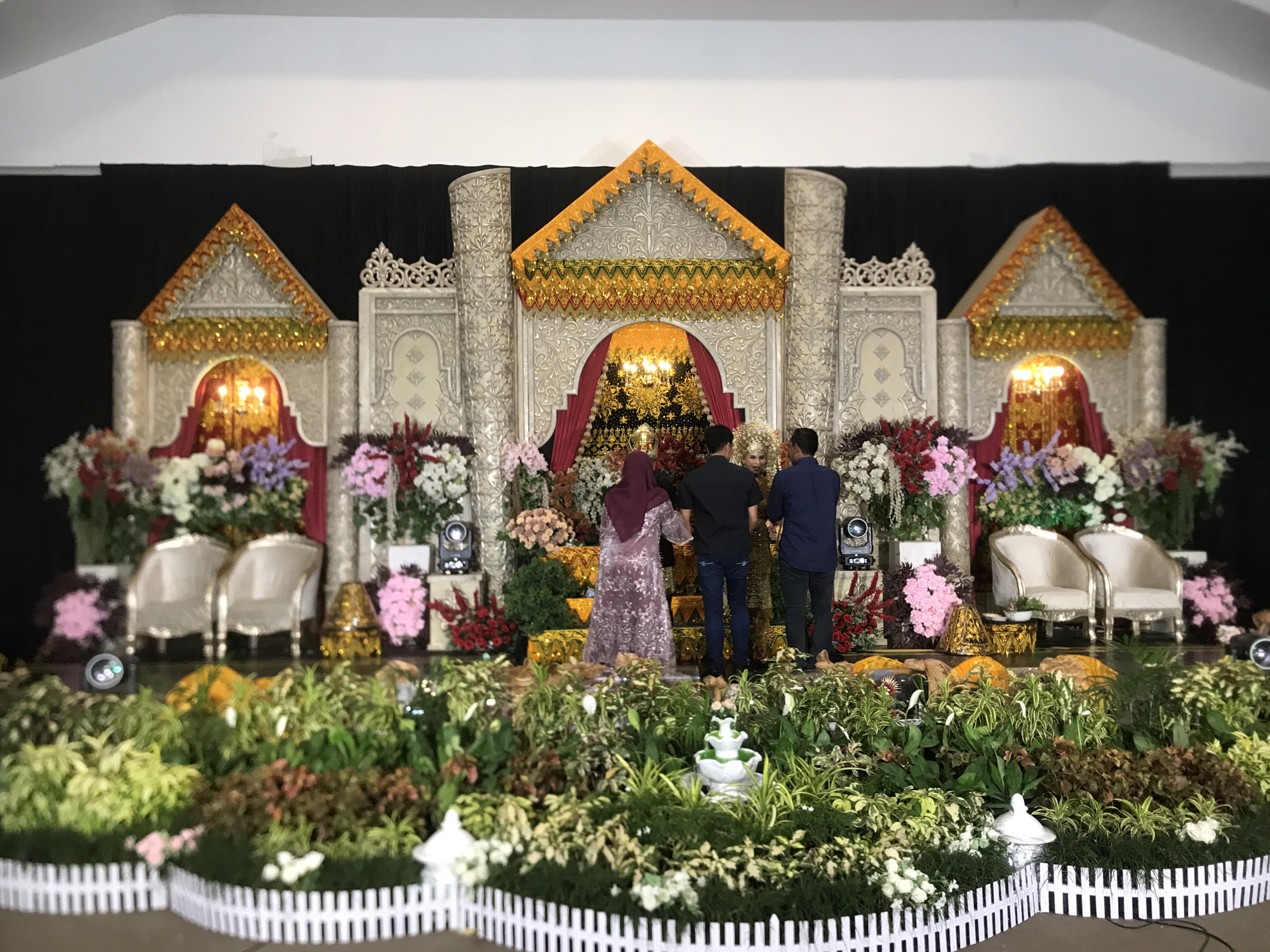 A wedding stage in Lhokseumawe, Indonesia is adorned with a central, ornately-decorated pavilion flanked by smaller, matching structures, all against a black backdrop. The stage includes two pairs of white chairs, each set accompanied by floral arrangements. A small fountain and potted plants line the base of the stage, bordered by a white picket fence. Three people stand on the stage facing away from the camera, two men in dark shirts and pants and a woman in a patterned dress and maroon headscarf. Lighting fixtures are visible near the floral arrangements.