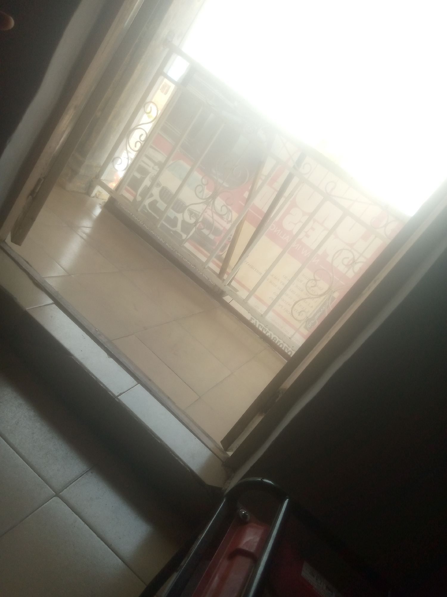 A wide-angle shot captures an interior view from a doorway looking out onto a tiled balcony. The lighting suggests it is daytime, with bright sunlight coming from an overhead area, creating a hazy effect and washing out details of the exterior.
The immediate foreground shows a dark, possibly wooden, threshold and the edge of a dark, matte surface on the right. In the center, a tiled floor extends from the doorway onto the balcony. The tiles are a neutral, light beige color and are laid in a grid pattern.
The balcony area is partially obscured by a metal railing with decorative scrollwork. Beyond the railing, indistinct shapes and colors suggest structures or objects, possibly a vehicle or signage, are present. There is text on a surface visible through the railing, but it is too blurred to read.
In the bottom right corner of the image, a red object with black metal framing is visible. It appears to be a piece of equipment or furniture, possibly a generator or a cart, with some visible fasteners.
The overall scene is characterized by strong light, some reflective surfaces on the tiled floor, and a sense of looking from a sheltered interior onto an open, bright exterior. The image composition is somewhat abstract due to the angle and lighting conditions.