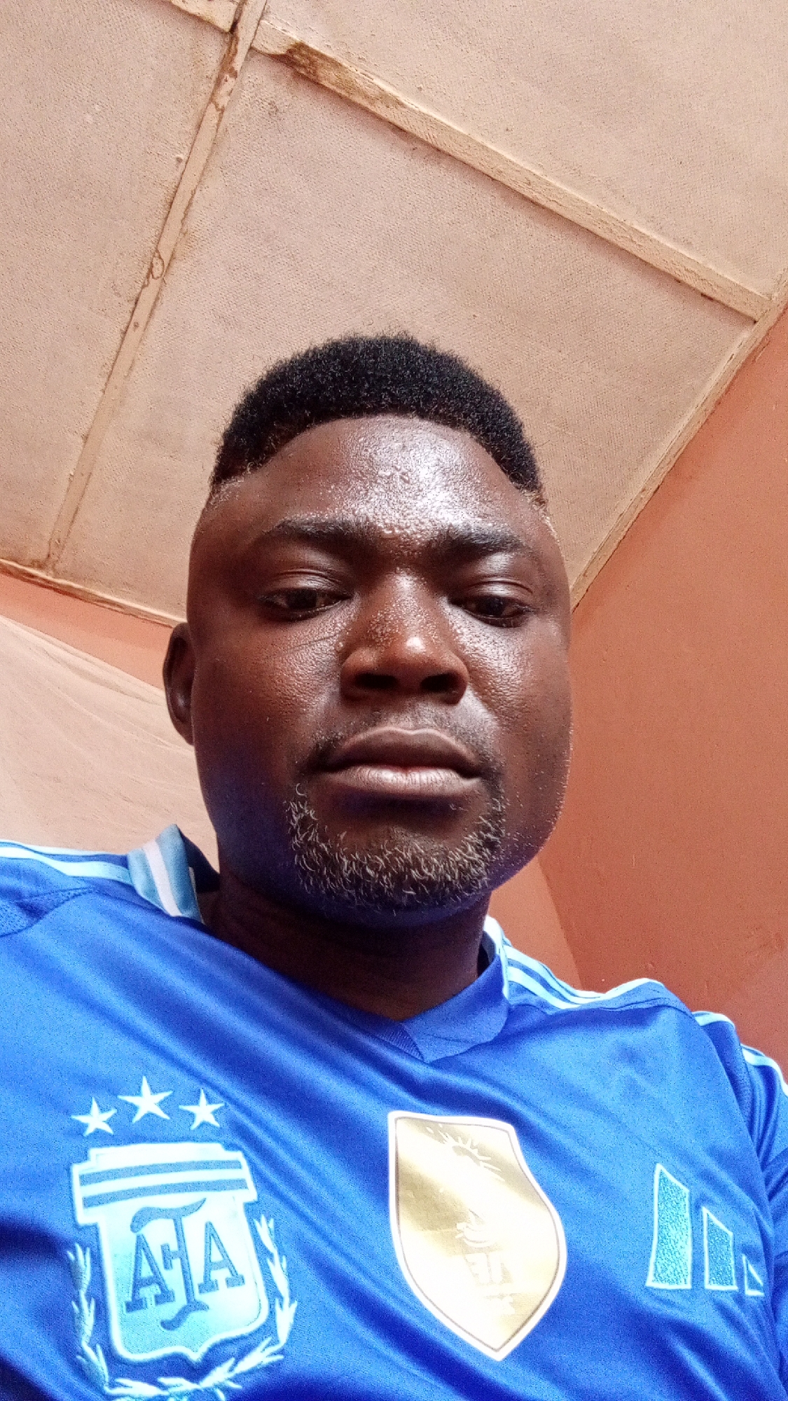 A close-up portrait shot features a man with dark skin, short dark curly hair, and a faded beard. He looks directly at the camera with a neutral expression. His forehead appears slightly sweaty or shiny.
He is wearing a vibrant blue short-sleeved sports jersey with light blue or white stripes visible on the shoulders, consistent with Adidas branding. On the left side of his chest, there is a light blue crest featuring the letters "AFA" (Argentine Football Association) encircled by a laurel wreath design, topped by three white stars. On the right side of his chest, a golden shield emblem, the FIFA World Cup Winners Badge, is prominent.
The background indicates an indoor setting. Directly above the man's head, a light brown or beige ceiling is visible, characterized by textured surfaces and what appear to be aged wooden planks or beams. To the man's right, a plain orange-peach colored wall fills the background. To his left, a darker reddish-brown wall is partly obscured by a thin, white mesh-like fabric or netting. The lighting is soft and diffuse, suggesting an indoor environment. No specific activity is depicted other than posing for the photograph. The visible text is "AFA".