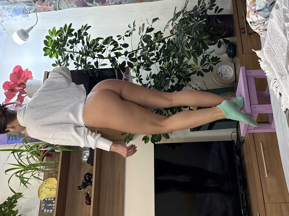 A woman in a white hoodie and patterned panties is lying prone with her legs extended on a small purple stool. She is wearing light green socks. Her body is positioned against a backdrop of lush greenery, including several potted plants and a leafy vine. In the background, there are a bookshelf, a television, and patterned wallpaper. The scene appears to be set in an indoor living space.