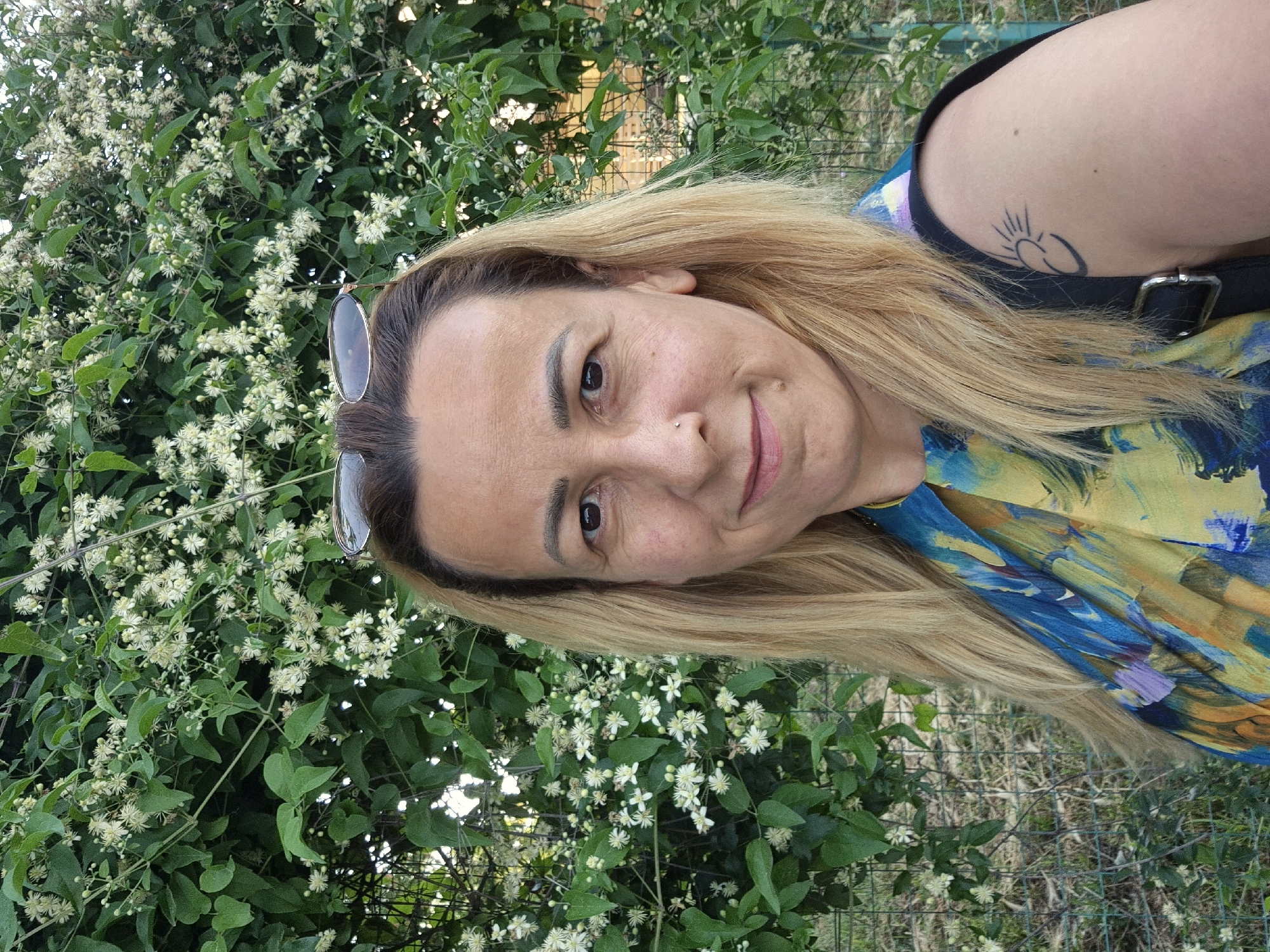 A medium shot of a woman with blonde hair, wearing a brightly colored shirt, stands in front of a bush with small white flowers. She is looking at the camera with a slight smile and has sunglasses on her head. Her left arm is visible, showing a tattoo of a sun. The background is a dense green bush with small white flowers and a glimpse of a fence. The lighting suggests it is daytime.