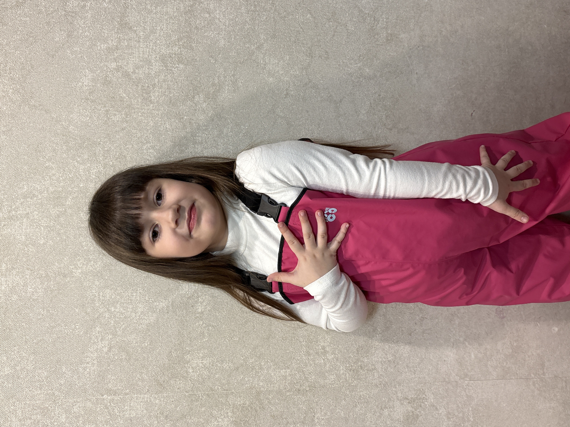 A young girl with long brown hair and bangs is lying on a light beige textured surface. She is wearing a white turtleneck shirt and pink snow pants with black suspender straps. Her hands are visible, one on her chest and the other behind her back. The background is plain and uniform. The exact location within Chaikovsky, Russia, cannot be determined from the image alone.