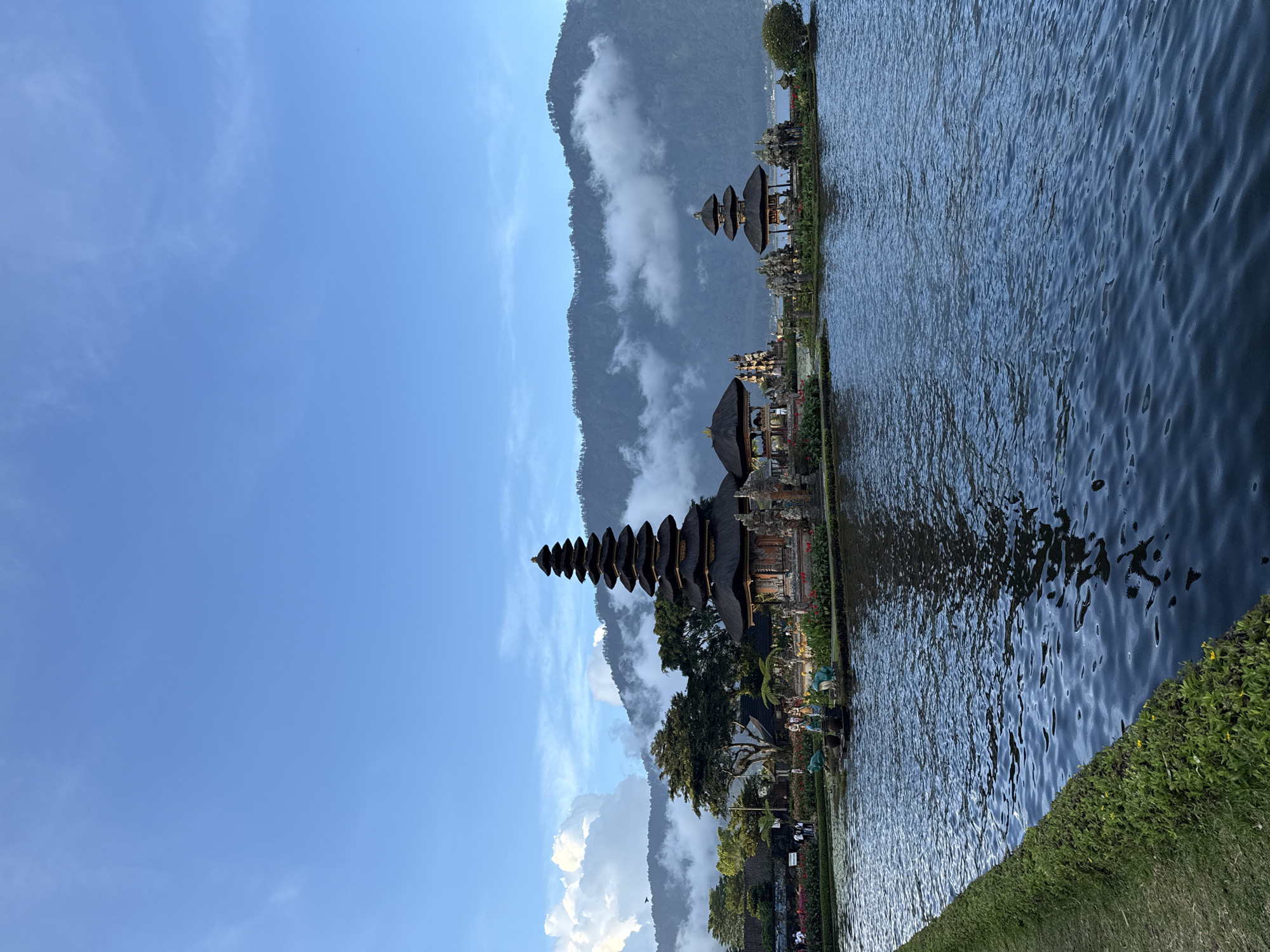 The image shows Pura Ulun Danu Beratan, a Hindu temple complex located on the edge of Lake Beratan in Baturiti, Bali, Indonesia. The temple, with its tiered pagodas, is partially obscured by trees and vegetation, and is reflected in the water. Mountains, shrouded in clouds, form the backdrop of the scene. The sky above is blue with some clouds. The foreground shows water and greenery along the edge of the lake.