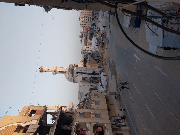 The image shows a scene of destruction in an urban environment. A mosque's minaret stands relatively intact amidst heavily damaged buildings. The street is clear but littered with debris. Several figures walk along the road. In the background, a row of intact buildings can be seen. The sky is clear. The image conveys a sense of conflict and devastation.