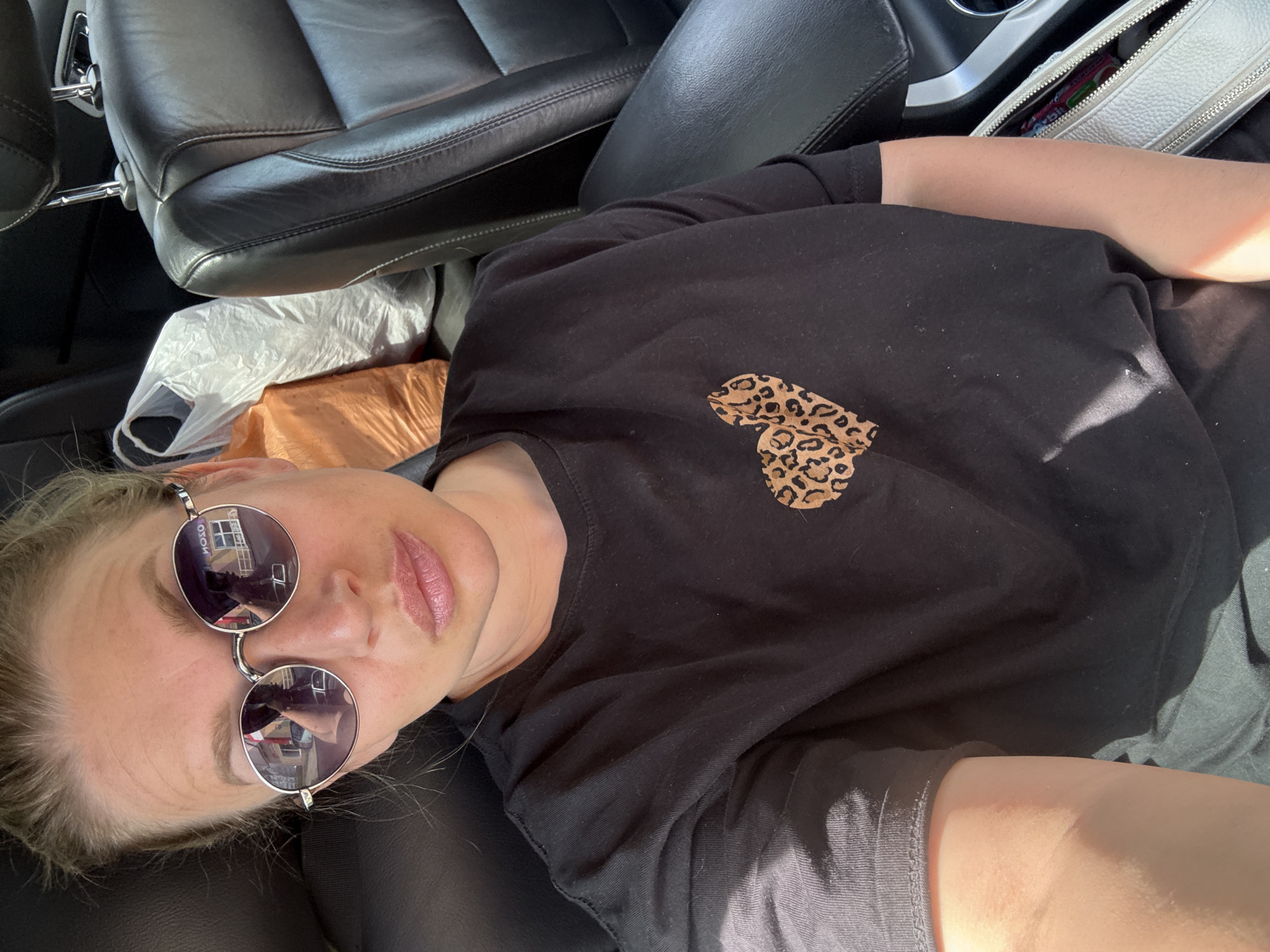 A medium shot captures a woman in a car, seated in the driver's seat, facing the camera. She is wearing a black t-shirt with a leopard print heart graphic on the left side of her chest and round, silver-rimmed sunglasses. Her blonde hair is pulled back. The interior of the car appears to be black leather. In the background, on the passenger side, are two plastic shopping bags, one orange and one white. The image was taken in the city of Valday, Russia.