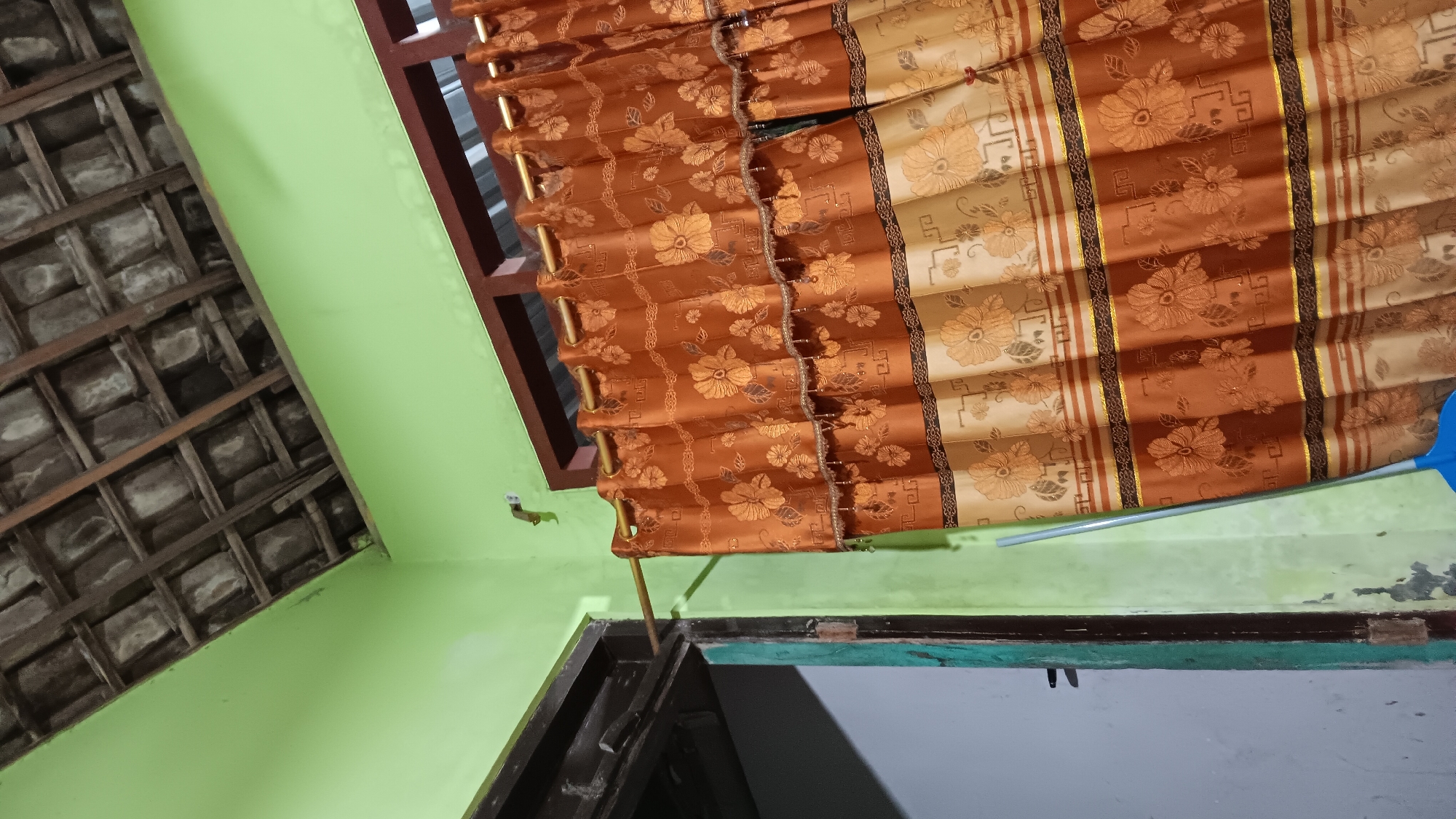 The image depicts an indoor scene, likely a room in a house located in Sonobekel, Indonesia, given the context. The foreground is dominated by a large, patterned curtain in shades of orange and gold, hanging from a brass rod. The curtain has a floral motif with decorative beaded trim. To the left, part of a doorway is visible, with a dark wooden frame and a glimpse of a grey wall beyond. A broom with a blue head leans against the wall to the right of the doorway and in front of the curtain. The upper part of the image reveals a section of the ceiling, which appears to be constructed from interlocking clay tiles supported by wooden beams. The walls are painted a light green color. The scene appears to be at dusk or indoors with artificial lighting, as the overall illumination is dim. There are no people visible in the image.