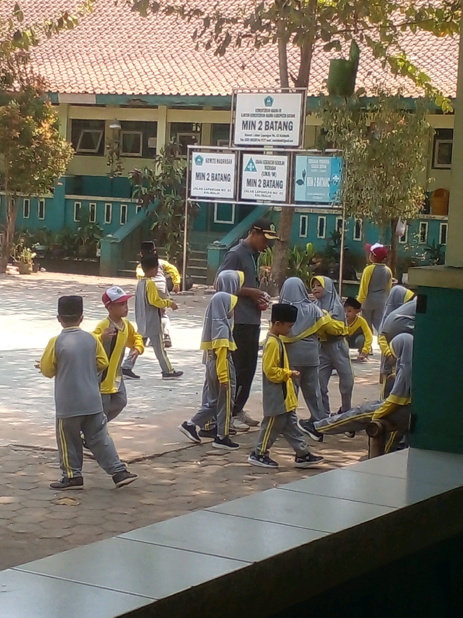 A group of children wearing school uniforms and a teacher are gathered in an outdoor courtyard. The children are dressed in grey pants and yellow long-sleeved shirts, with some also wearing grey hijabs. The teacher is wearing a grey shirt and a dark cap. The courtyard appears to be part of a school, with a building in the background that has a tiled roof and a row of windows. Signs indicating "MIN 2 BATANG" are visible, suggesting the location is Madrasah Ibtidaiyah Negeri 2 Batang. One sign, in particular, lists the address as "JALAN LAPANGAN NO. 03 KALIBARU." Banyuputih, Indonesia, is the identified city. The children are interacting, with some standing, others walking, and one child appears to be crouching. The ground is paved with interlocking stones, and sunlight casts shadows.
