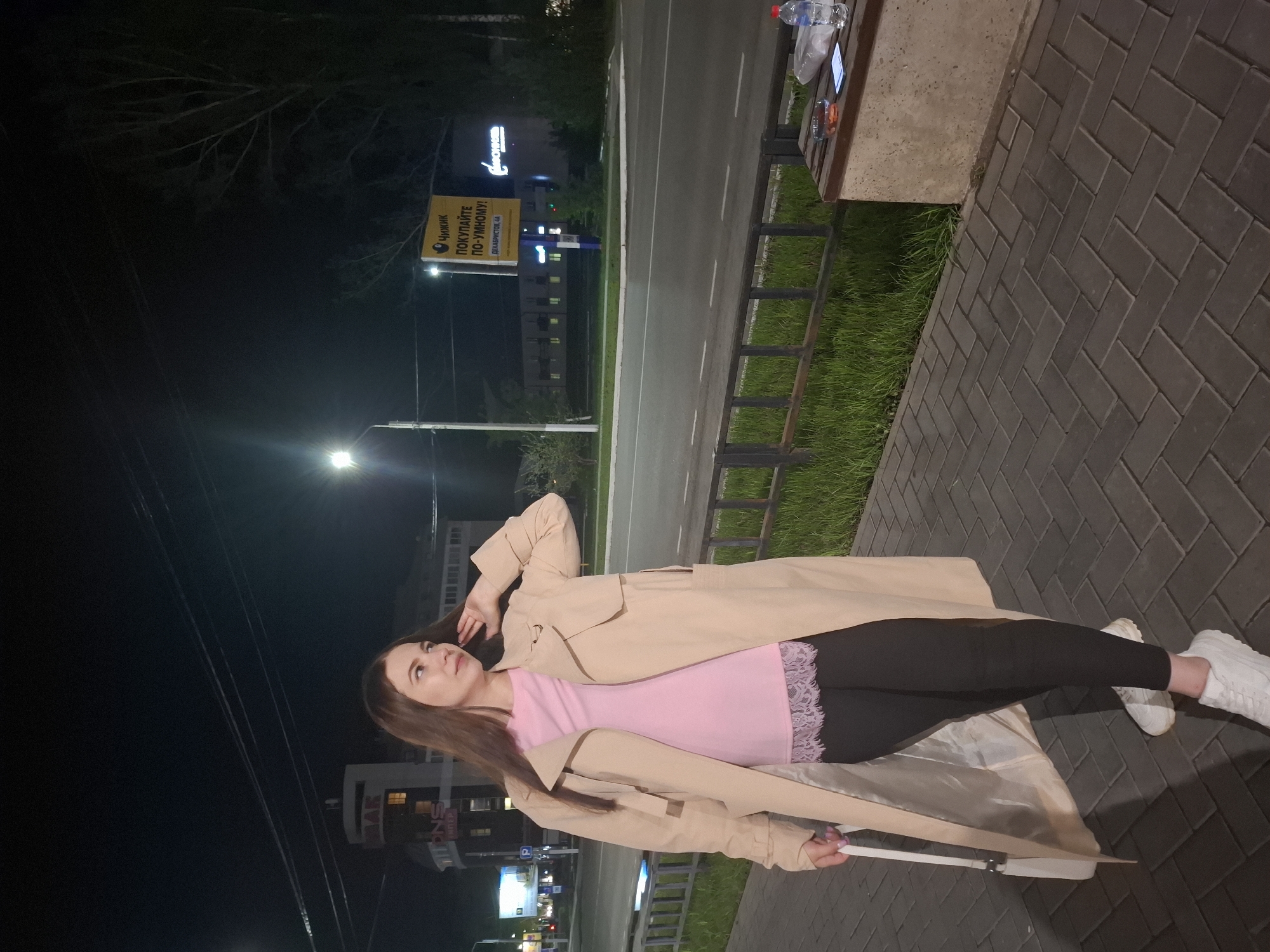 A young woman is lying on a bench at night in the city of Chaikovsky, Russia. The location is discernible by the "Chizhik" advertising sign on a pole visible in the background and the text "ДЕКАБРИСТОВ, 44" (Dekabristov, 44) which refers to the address of a location in Chaikovsky. The woman is wearing a pink top with lace trim, black leggings, a light-colored trench coat, and white sneakers. She is holding a white purse or bag with a thin handle. She is lying on her side with her head propped up by her hand. A paved walkway is visible, along with a street, a bench, and some vegetation. Other signs, lighting, and buildings are visible in the background. There is a water bottle and what appears to be an electronic device on the bench. The sky is dark, and a street light illuminates the scene.