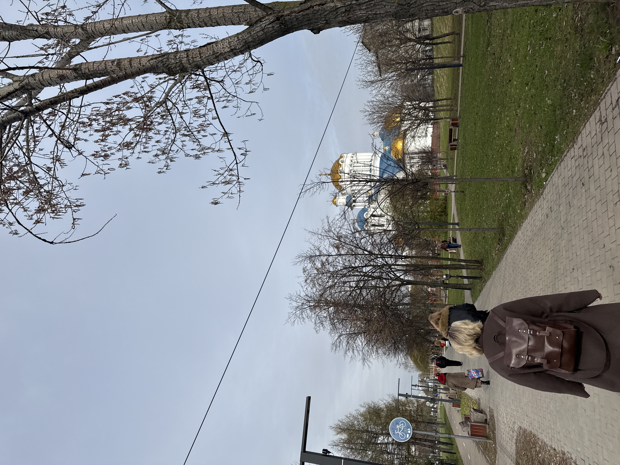 A medium shot captures a scene in Moscow, Russia, on a paved path. The image is rotated slightly counter-clockwise. A woman with a blonde bob hairstyle and a brown coat, carrying a brown leather backpack, walks away from the viewer. Other pedestrians are visible in the background. The Church of Christ the Savior with its gold domes is visible between bare trees. Bare tree branches also frame the top left corner of the image. A power line cuts across the sky. A sign depicting a bicycle icon is present in the lower left. The weather appears overcast. A green lawn and additional trees are adjacent to the paved path.