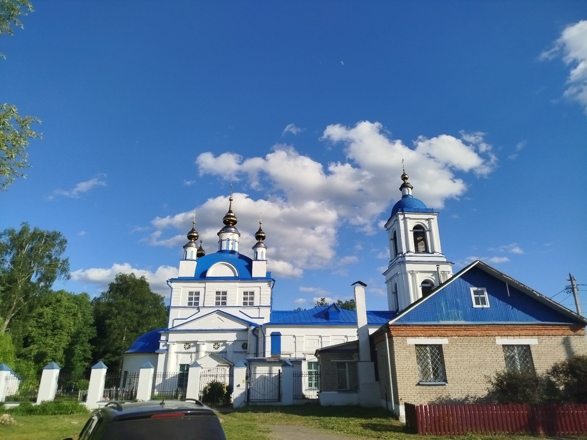 The image shows a church in Мамонтово, Russia, under a blue sky with scattered clouds. The church is predominantly white with blue roofs and domes topped with golden crosses. A bell tower rises on the right side of the main structure. A brick building with a blue roof is adjacent to the church on the right. A red fence surrounds the brick building. A black vehicle is partially visible in the foreground. Trees and greenery surround the structures.