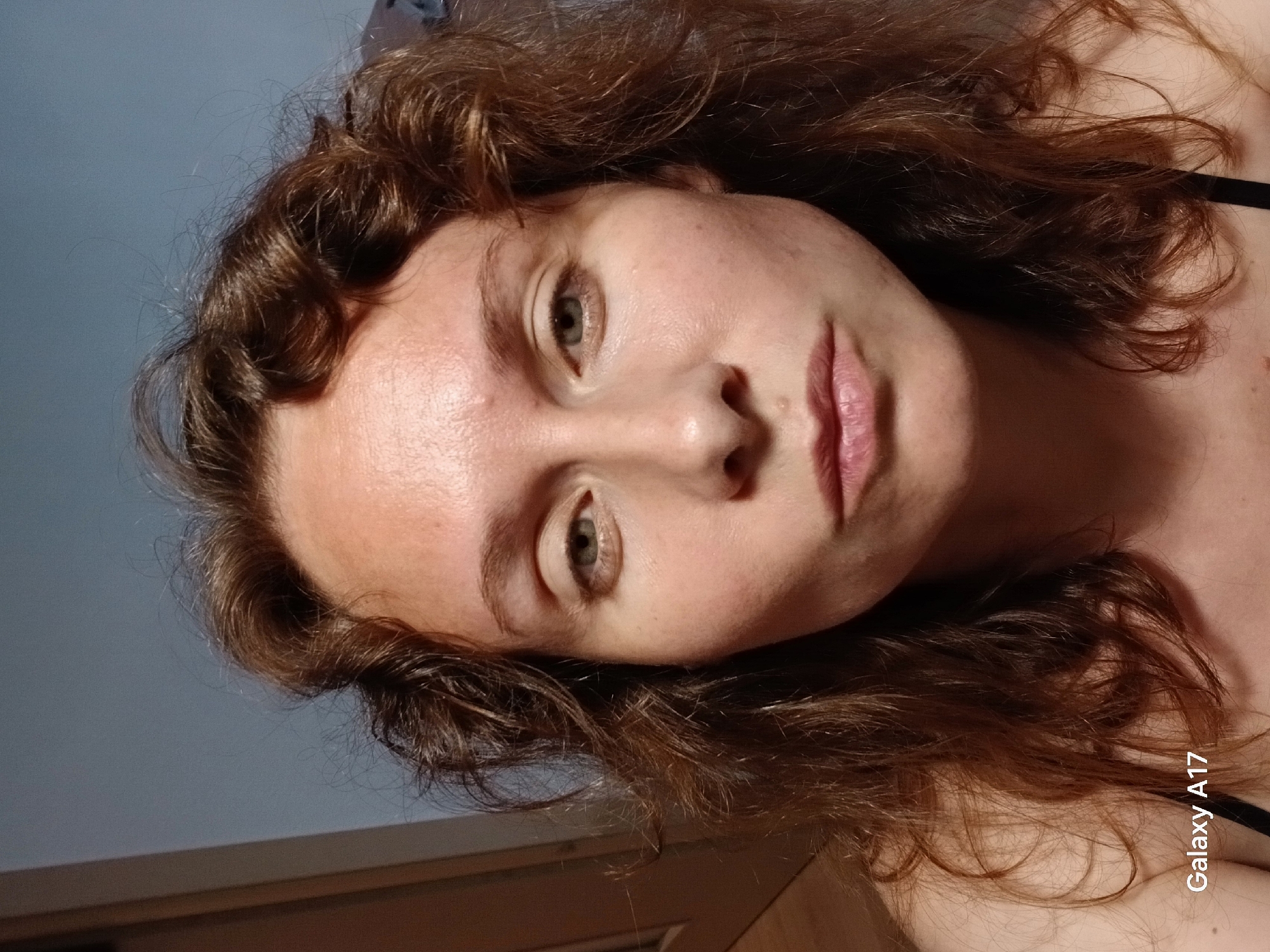 This is a close-up portrait of a young woman, likely taken indoors in Yekaterinburg, Russia. Her head is tilted to her right (the viewer's left), and she gazes directly at the camera with a neutral and calm expression.
The woman has light-colored eyes, possibly green or hazel, with prominent pupils. Her eyebrows are natural and well-defined. Her skin is fair, showing some natural redness, minor blemishes, and a few small moles or freckles on her forehead, cheeks, and chin. Her lips are naturally pink and slightly parted.
Her hair is a medium to dark brown with warm, reddish undertones, and features a distinct curly or wavy texture. It frames her face and extends out of view to the sides and bottom of the frame. A dark strap, possibly from a piece of clothing, is visible on her right side (image left).
The background is minimalist, consisting of a plain, light blue or grey wall in the upper left portion of the image. A dark, horizontal line, possibly a piece of furniture or architectural trim, is visible across the bottom of the frame, leading to a darker, shadowed area. The lighting is soft and even, suggesting ambient indoor light.
The text "Galaxy A17" is visible vertically in the bottom right corner of the original image, indicating the device used for capture. The image is presented with a 90-degree clockwise rotation relative to a typical upright portrait.