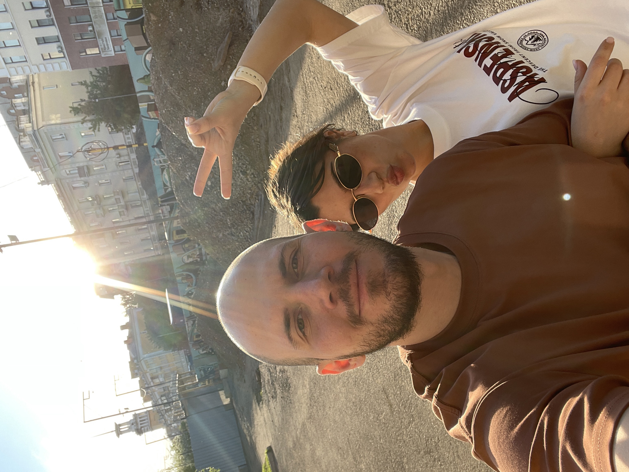 The image captures a casual outdoor photo of two individuals in an urban setting, identified as Krasnoyarsk, Russia. The sun is low in the sky, creating a strong lens flare on the left side and casting a warm, golden light across the scene, indicative of late afternoon or early evening. The weather is clear and sunny.
In the foreground, a man with a short beard and a bald head is visible, wearing a plain brown t-shirt. He is looking directly at the camera with a subtle smile. Behind him and slightly to his right is another person with dark hair, wearing dark, round sunglasses and a white t-shirt. This individual is making a "peace" sign with their left hand, which also features a white wristband. Their face is partially obscured by the sunglasses and a slight pout.
The activity suggests a relaxed moment, possibly a selfie taken by the man in the foreground. The background features several multi-story buildings, consistent with a city environment. On the white t-shirt worn by the person in the background, partial text is visible, reading "THE DESCENT FROM SNOW VALL..." and "ASPEN...", along with a circular graphic. On a distant building in the background, some Cyrillic text can be faintly discerned as "ПЕРВЫЙ ЦЕНТР..." (First Center...). The ground appears to be paved, like a street or pathway.