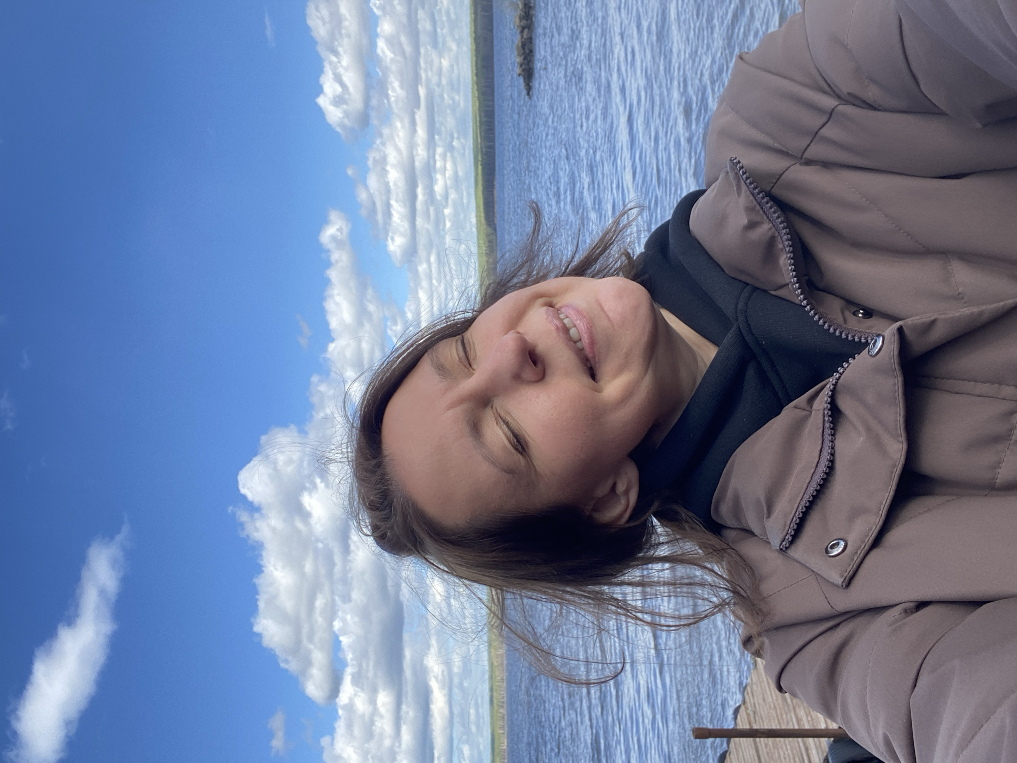 A woman is smiling in a close-up outdoor portrait shot at the shore of a body of water, possibly a lake or river. She is wearing a brown puffer jacket over a black hoodie. The sky is blue with fluffy white clouds, and a shoreline with vegetation can be seen in the distance across the water. The location may be the city of Chaikovsky, Russia.