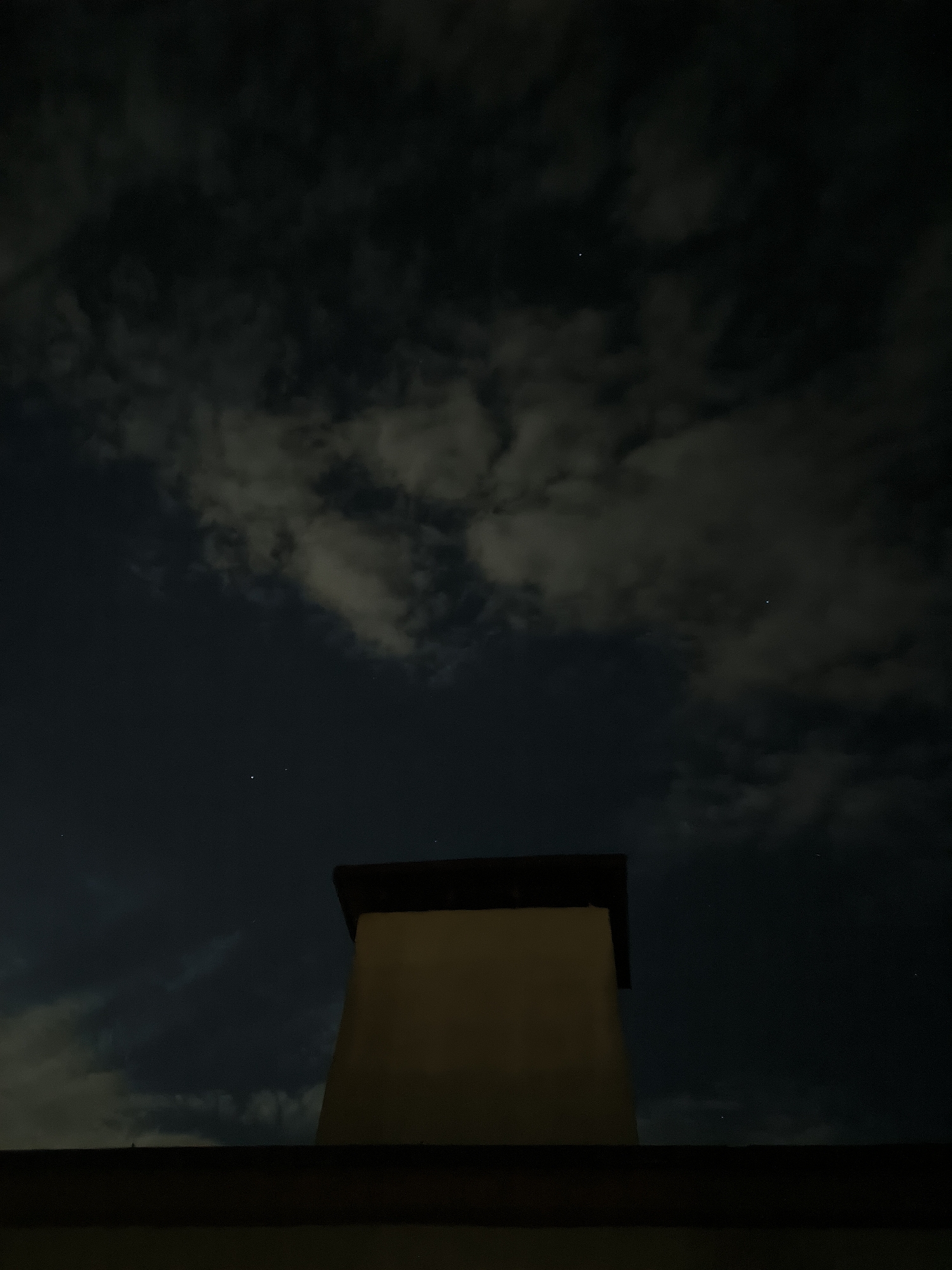 The image depicts a nighttime scene. In the foreground, the top of a building and a chimney are visible against a dark sky filled with clouds. The sky is partially illuminated, with some areas appearing brighter due to the clouds reflecting light. A few faint stars are visible in the sky. The scene is devoid of any people or discernible activity. The overall impression is one of a quiet, urban night. There is no information within the image to suggest a specific location within Chișinău, Moldova.