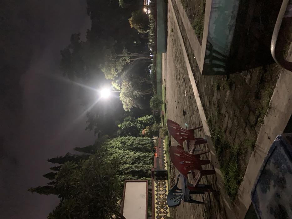 The image captures an outdoor scene at night, likely in Metro, Indonesia, illuminated primarily by a bright street lamp. The setting appears to be a public space such as a park, plaza, or garden, featuring a paved pathway or ground surface bordered by low concrete structures. Lush green trees and dense foliage dominate the background and mid-ground, with their leaves appearing bright under the artificial light.
In the foreground and mid-ground, several plastic chairs are visible. Two red chairs are prominently placed, and a blue chair is partially visible near the bottom left, next to a decorative wall with an intricate geometric pattern. Another blue chair is cut off by the right edge of the frame. A rectangular white board or sign stands near the patterned wall, but no text is discernible on it. The ground, a mix of paved sections and patches of dirt and grass, shows some scattered debris or leaves.
The sky is dark, suggesting overcast conditions that diffuse the light from the lamp, creating a hazy glow around it. No people are present, lending a quiet and still atmosphere to the scene. There is no specific activity taking place, though the arrangement of chairs implies a gathering spot that is currently empty.