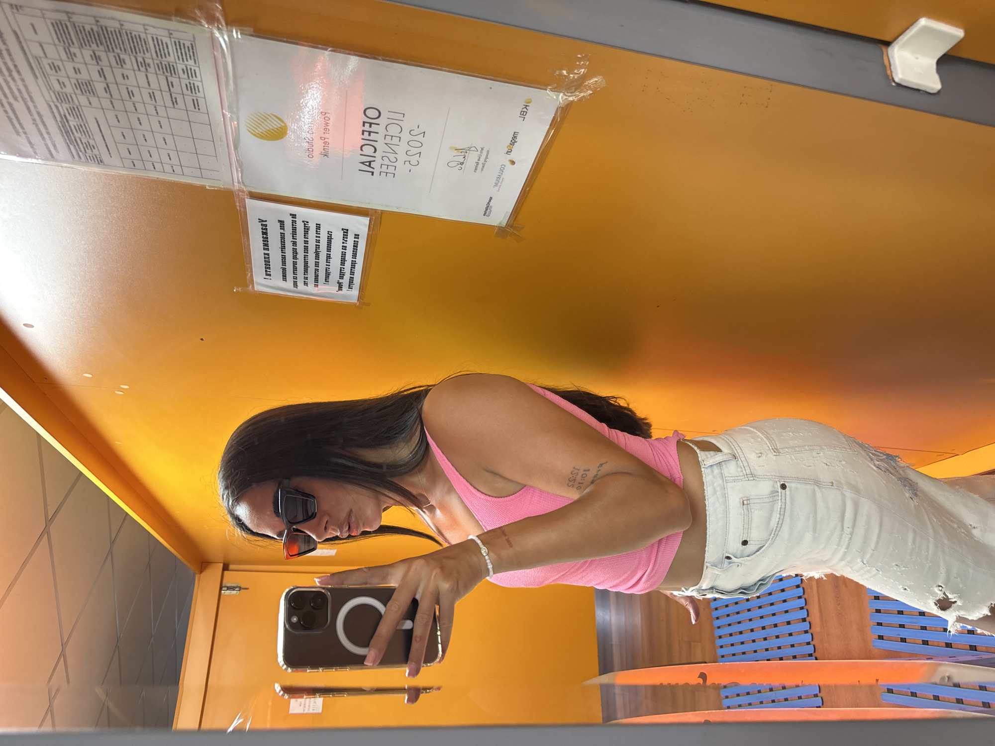 This is a selfie taken by a young woman in Pernik, Bulgaria. She is wearing a pink top and distressed white jeans, and has on large, dark-rimmed sunglasses. She is holding her phone, which has a clear case with a white ring on the back, to take the picture. The background is a bright orange wall with some signage. The signage includes text in what appears to be Bulgarian, with some English words visible, such as "OFFICIAL LICENSE" and "Power Bank". The overall scene appears to be indoors, possibly in a public space or a waiting area, given the seating visible in the reflection. The lighting suggests it could be daytime. The woman's expression is neutral, focused on taking the selfie. There are some visible tattoos on her arm.