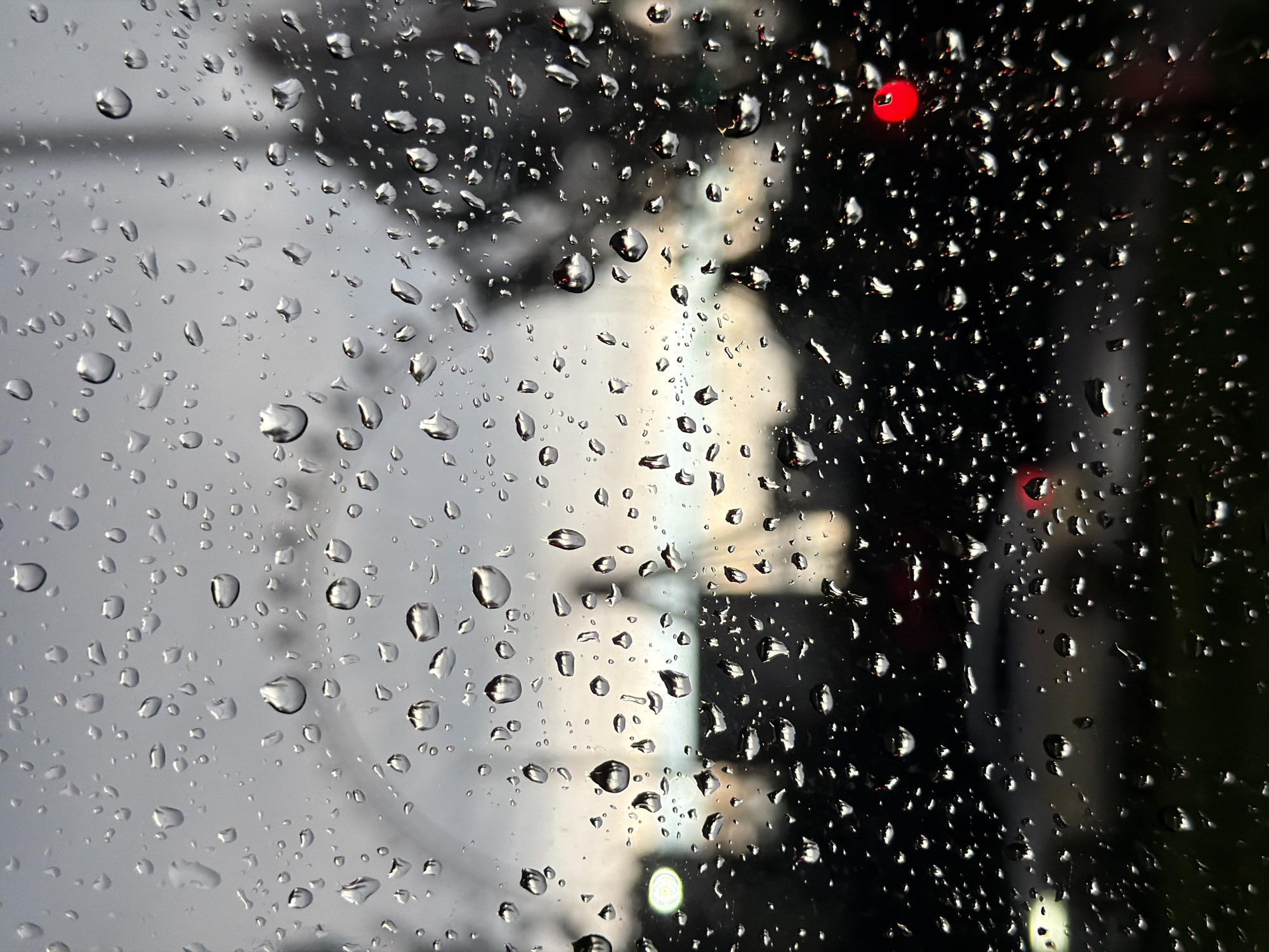 The image depicts a rainy day in Bishkek City, Kyrgyzstan, viewed through a rain-covered window. The focus is on the water droplets clinging to the glass, obscuring and refracting the scene outside. The background shows indistinct shapes of buildings, suggesting an urban environment. Red and white lights are also visible in the background, possibly from vehicles or streetlights. The overall tone of the image is moody and blurred, emphasizing the atmospheric effect of the rain.