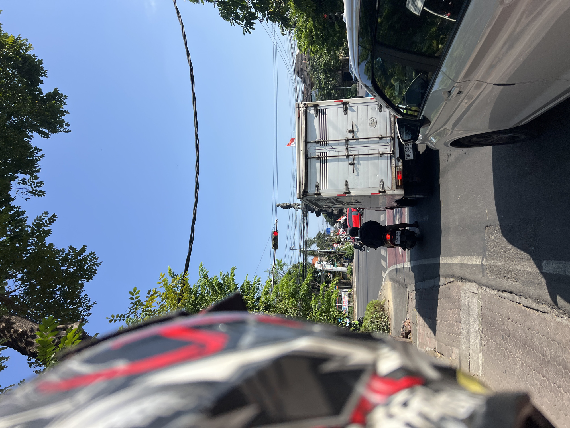 The image captures a street scene in Denpasar, Indonesia, likely during the daytime due to the bright sunlight. In the foreground, the left side of a motorcycle helmet, with red and white accents, is visible, suggesting the photo is taken from the perspective of a rider.
The scene depicts a street with multiple vehicles. A white box truck is in the center of the frame, facing away from the viewer. It has a small Indonesian flag on its roof. To the right of the truck, the side of a silver car is visible, also facing away. Behind the truck, a person on a scooter, wearing a black jacket and black helmet, is also facing away and appears to be waiting or moving slowly in traffic. Further down the road, other vehicles and a red car can be seen.
Traffic lights are visible, indicating an intersection. One traffic light is red. Trees and power lines are present along the street, with a clear blue sky above. Buildings and some signage are visible in the background, suggesting an urban environment. The road surface is asphalt with painted white lines. The sidewalk area has a textured paving.