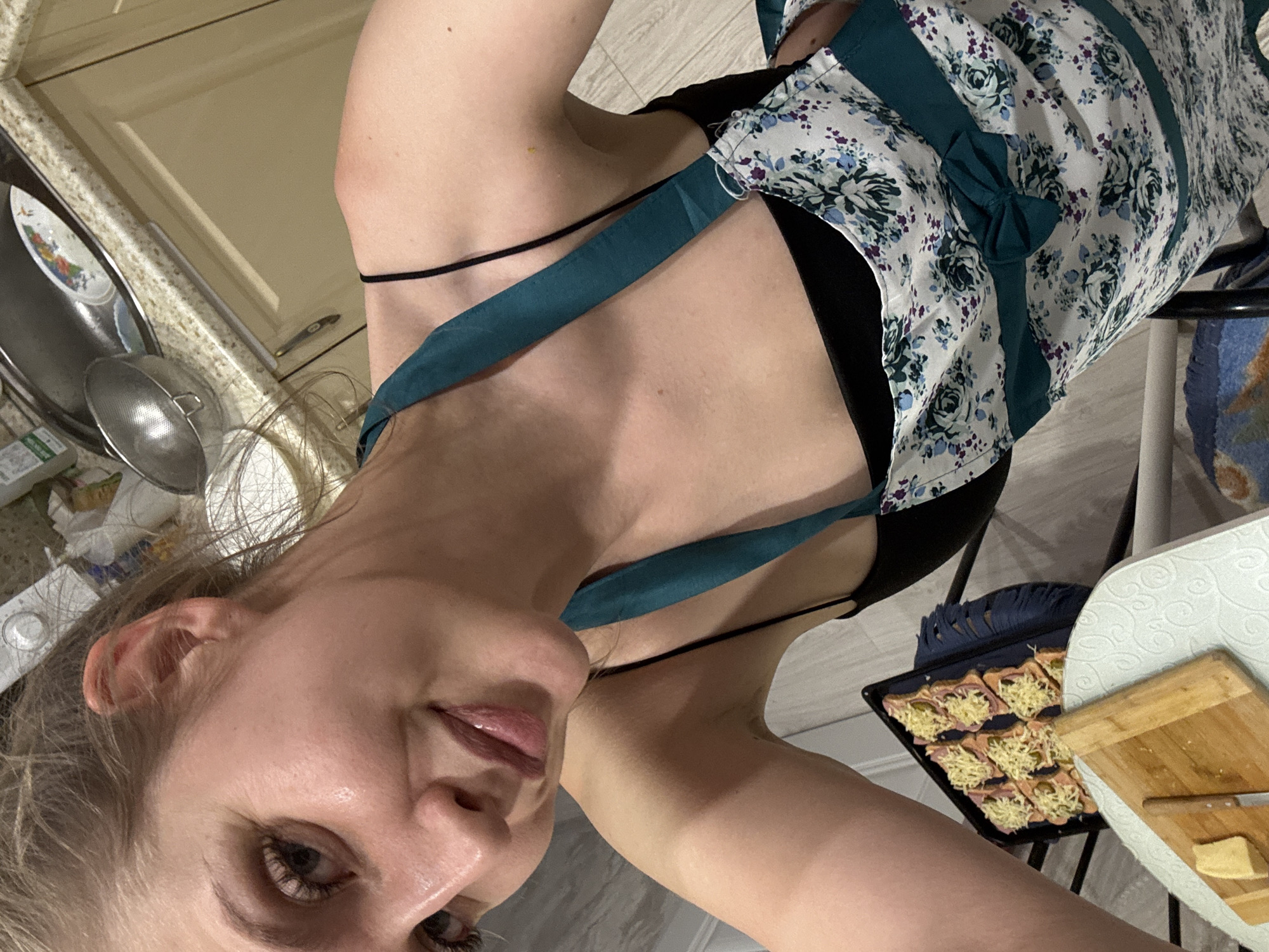 A person with fair skin and blonde hair is in a kitchen, wearing a floral apron with green trim and a black top. They are facing the camera at an angle. In the background, there are kitchen cabinets and a countertop with a sink and various utensils. A baking sheet with small sandwiches topped with cheese and a wooden cutting board with a knife and small pieces of food are visible on a surface near the person. The location cannot be determined to be in Izhevsk, Russia from the image alone.