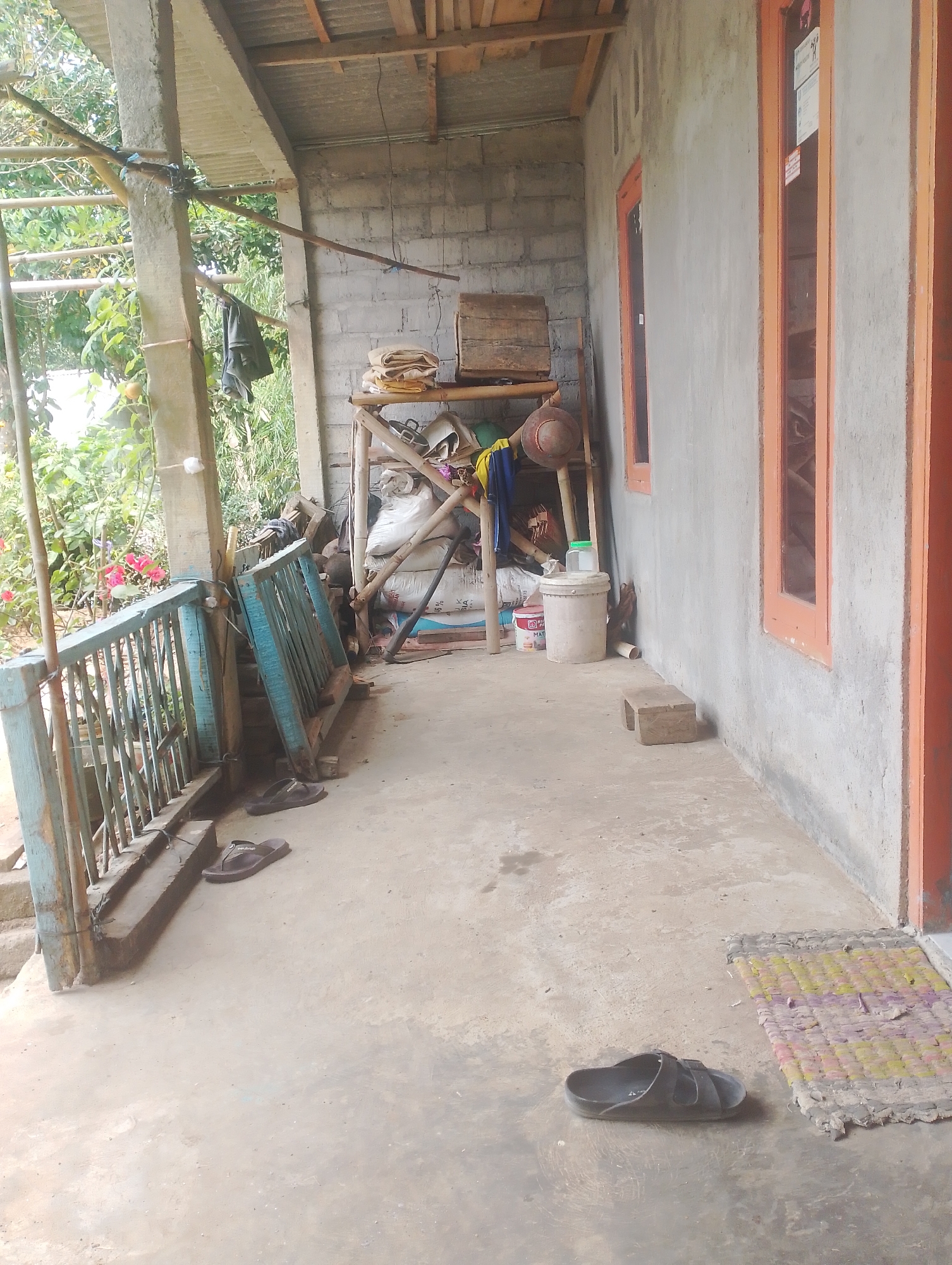 The image depicts the exterior of a building in Krasak, Indonesia. The scene is an outdoor porch area. On the left side of the image, there is a blue wooden railing. In front of the railing, a pair of dark brown flip-flops are placed on the concrete floor. Further to the left, another pair of similar flip-flops are also visible.
The building itself has a grey concrete wall with two orange-framed windows. The roof of the porch is constructed from corrugated metal supported by wooden beams.
In the background, there is a makeshift shelving unit made of bamboo and wood, piled with various items. These include sacks, what appears to be fabric, and other miscellaneous objects. A bucket with a green lid is also present. To the right of the shelving unit, a small, square wooden stool is positioned on the floor near the wall. A worn, colorful doormat lies on the ground in the lower right corner of the image. Green foliage and some pink flowers are visible behind the railing on the left. The ground is a plain concrete surface. There are no people visible in the image.