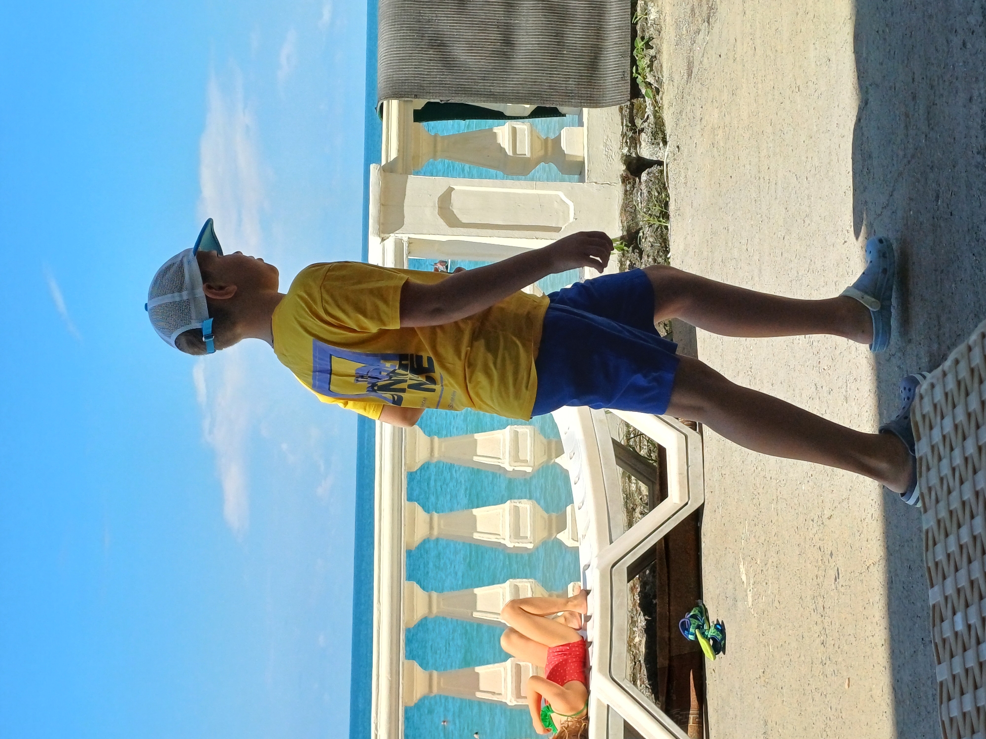 In Sochi, Russia, a young boy wearing a yellow t-shirt and blue shorts walks along a sunlit promenade with the Black Sea in the background. He is looking to his right, away from the camera. Further down the promenade, a young girl lies on a white lounge chair, wearing a red swimsuit and holding a green object. The sea is a bright turquoise, and a white railing with ornate pillars separates the promenade from the water. A striped towel hangs on the railing to the right of the boy. The ground of the promenade is paved with concrete, and there are some rocks and sparse vegetation at the edge. The sky is clear and blue.