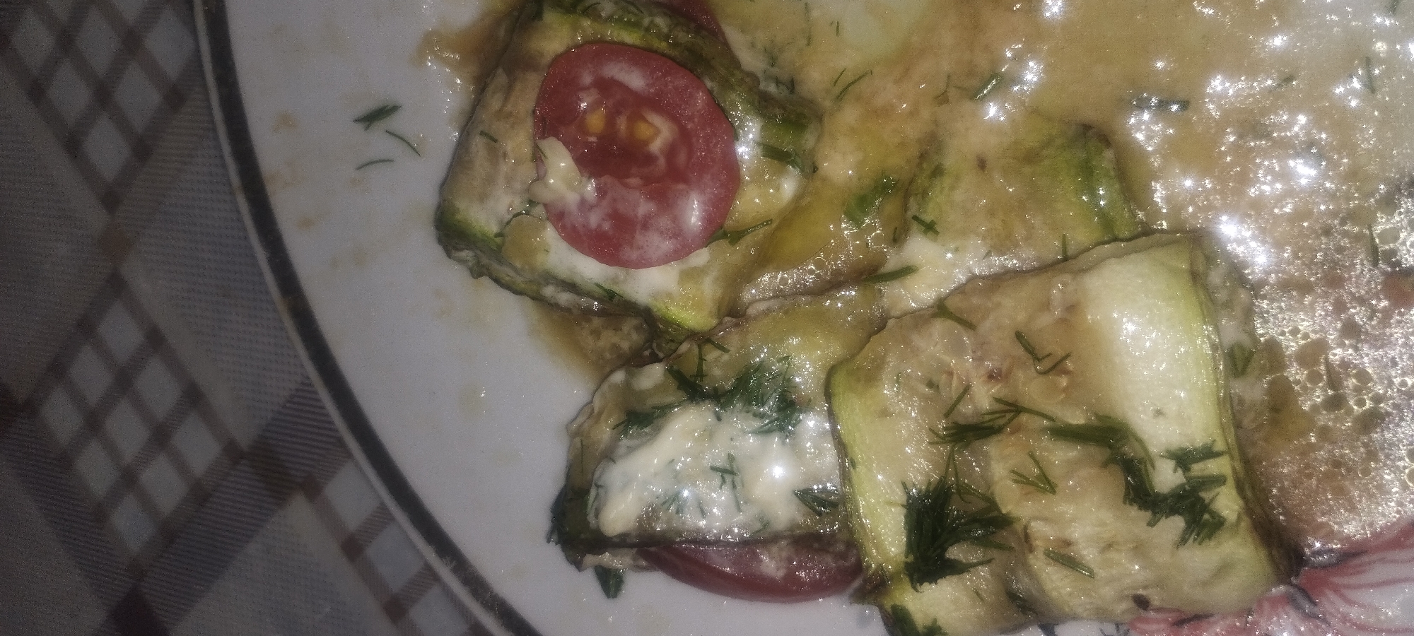 The image shows a close-up view of a dish of food, likely zucchini rolls, served on a plate. The dish consists of slices of zucchini wrapped around a filling and topped with a creamy sauce and chopped dill. There are also slices of tomato visible on top of some of the zucchini rolls. The food appears to be glistening, indicating it may be saucy or oily. The plate is white with a dark rim and is placed on a checkered tablecloth. The lighting in the image is dim, with some reflections visible on the surface of the food, suggesting a flash was used. The background is out of focus, showing the pattern of the tablecloth. There are no people or any indication of a specific location within a city.