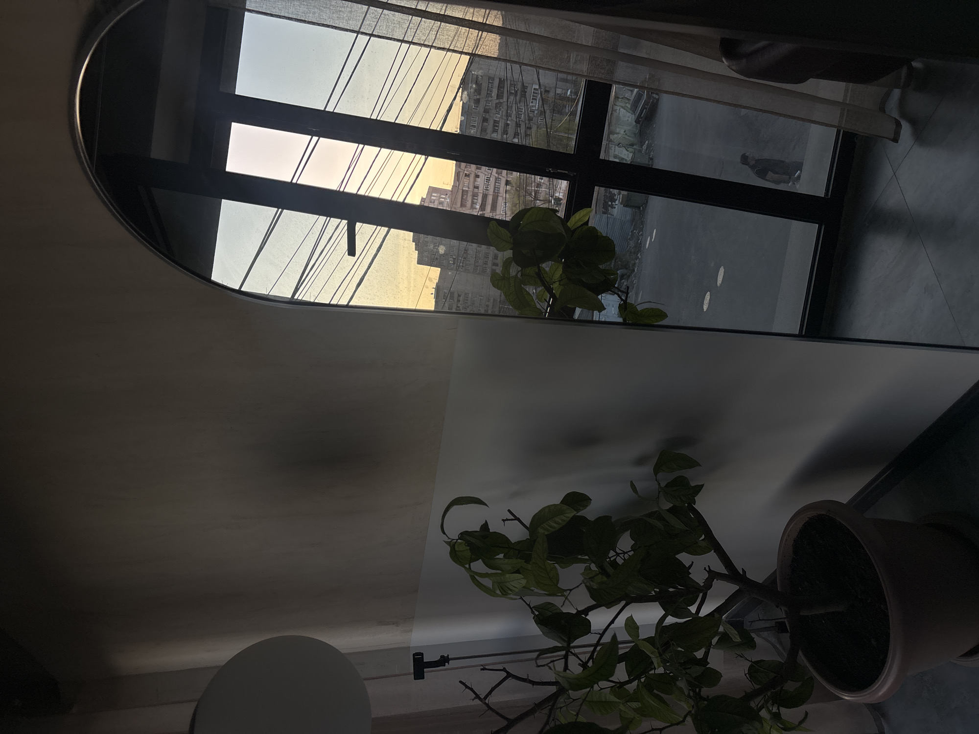 An interior view through a window in Yerevan, Armenia.
Through a dark-framed window, a building exterior is visible during what appears to be either sunrise or sunset, with golden light diffused through the sky and the tall apartment buildings across the street. Power lines are visible as thin black lines against the bright sky. The area visible outside the window appears to be a street or sidewalk. A pedestrian is walking.
Inside, a potted plant with green leaves sits on the ledge in front of the window. The wall appears to be painted in two tones: a lighter color in the upper portion, and a darker shade lower down.