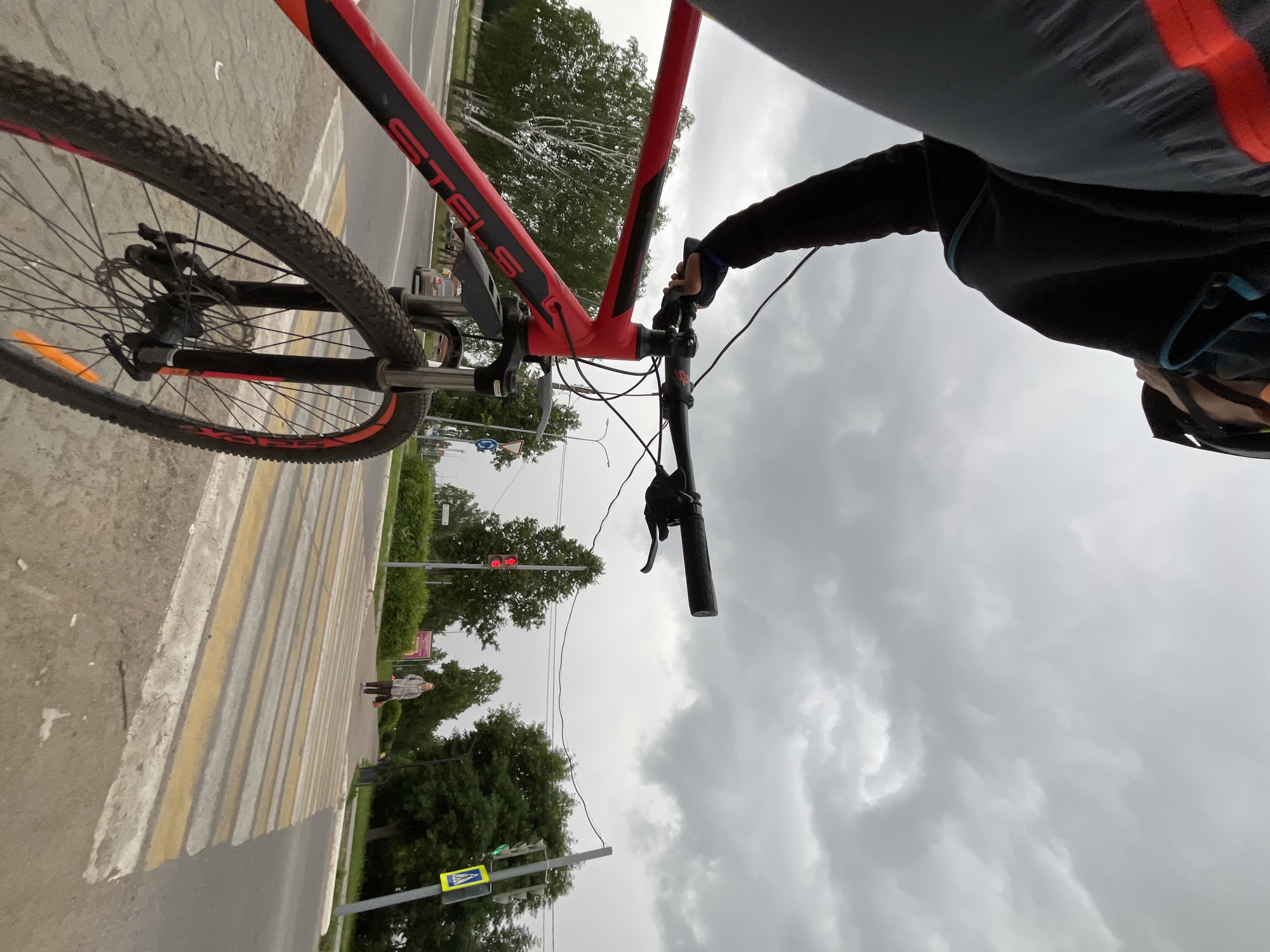 An overcast day in Chaikovsky, Russia, with a person on a red and black bicycle at a pedestrian crossing. The cyclist, wearing a black jacket, grey pants with a red stripe, and a helmet, is holding the handlebars. The bicycle's front wheel, with knobby tires and disc brakes, is in the foreground. In the background, a person walks across the street, carrying a red bag. Traffic lights are visible, with one showing red and another green. Trees line the street, and storm clouds gather in the sky. The location is likely near a road intersection, with a marked pedestrian crossing.