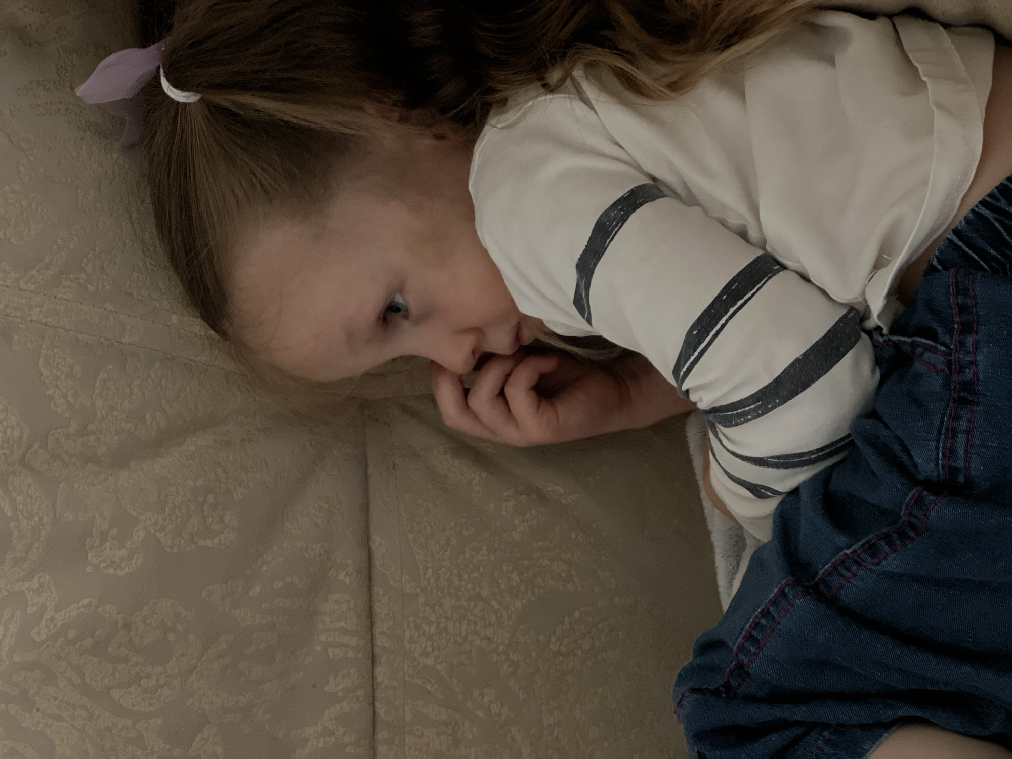 A young child with light skin and blue eyes is lying on a patterned beige surface, possibly a couch or bed. The child has blonde hair with a purple hair tie holding a small ponytail. They are wearing a white and gray striped long-sleeved shirt and blue denim pants. The child is facing left and appears to be resting their head on the surface, with their fingers near their mouth. The scene appears to be indoors, and the location is Unknown, Unknown.