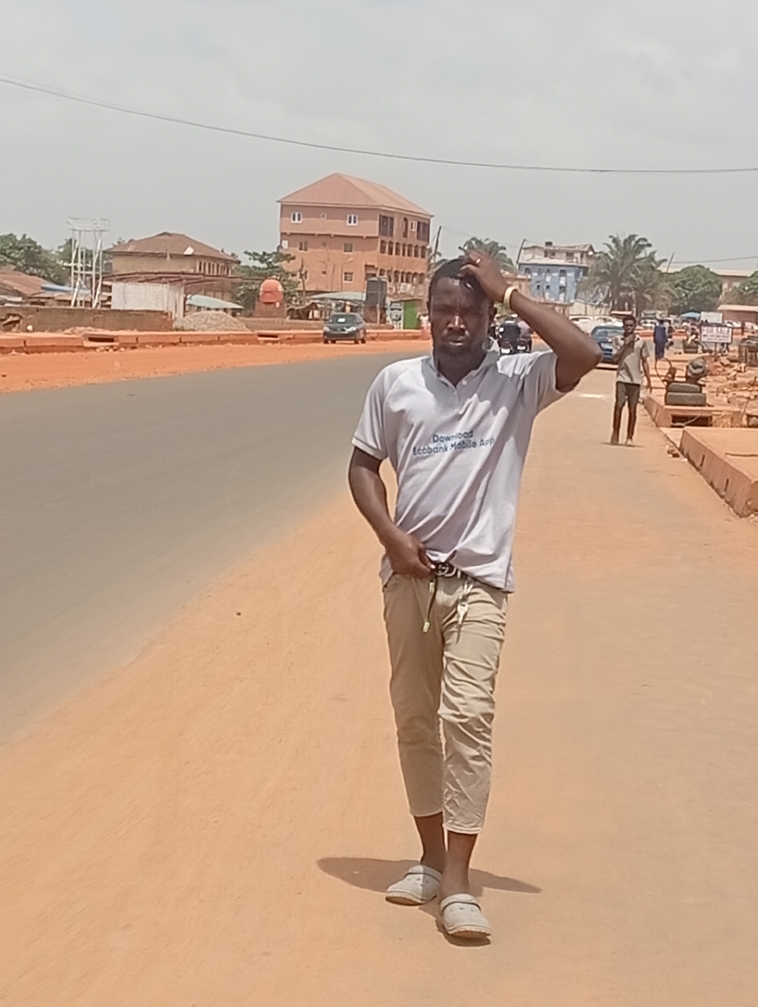 The image captures a full-body shot of a man standing on an unpaved roadside in what appears to be a town or city, identified as Yelwa, Nigeria. The man is positioned slightly to the right of the frame, looking directly towards the camera with a neutral expression. His right hand is raised, touching his hair, while his left hand rests near his waist, possibly in a pocket or holding onto his belt.
He is wearing a light grey short-sleeved polo shirt, light khaki or tan cropped pants, and light-colored slip-on shoes or sandals. Visible text on his shirt reads "Download Ecobank Mobile App". Keys are hanging from his belt on the left side. He casts a shadow to his right, suggesting the sun is high and to his left, indicating it is daytime.
Behind the man, a paved road extends into the distance, with an unpaved, reddish-brown shoulder on both sides. In the background, various buildings are visible, ranging from single-story structures to multi-story ones with red or brown roofs. One building on the left features a tall metal water tower-like structure. Piles of sand or gravel suggest ongoing construction or development. Several cars are parked or moving on the road in the mid-ground. Another man in a light shirt is seen walking further down the road on the right, and faint indications of a "FOR SALE" sign are visible near the right edge. The sky is light and hazy, characteristic of a sunny day. The overall scene depicts a typical street environment in a bustling, semi-urban setting.