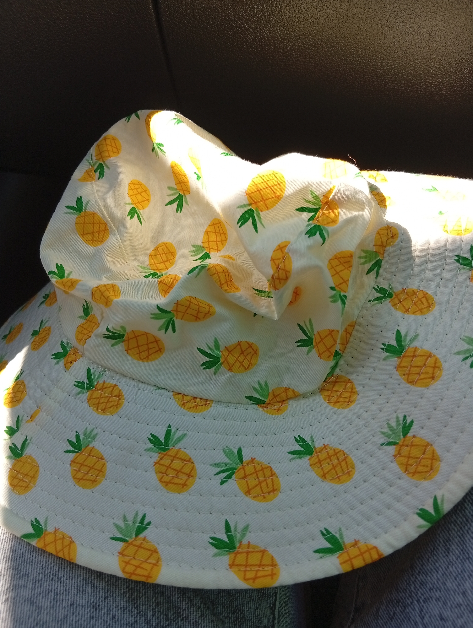 A white bucket hat with a yellow pineapple pattern is resting on a person's lap. The hat is made of fabric and has a wide brim. The pineapples are depicted with yellow bodies and green leaves, and they are arranged in a repeating pattern across the hat. The hat is partially illuminated by sunlight, which casts shadows on the fabric. The background is dark and appears to be the interior of a car. The bottom of the image shows the top of a pair of blue jeans.