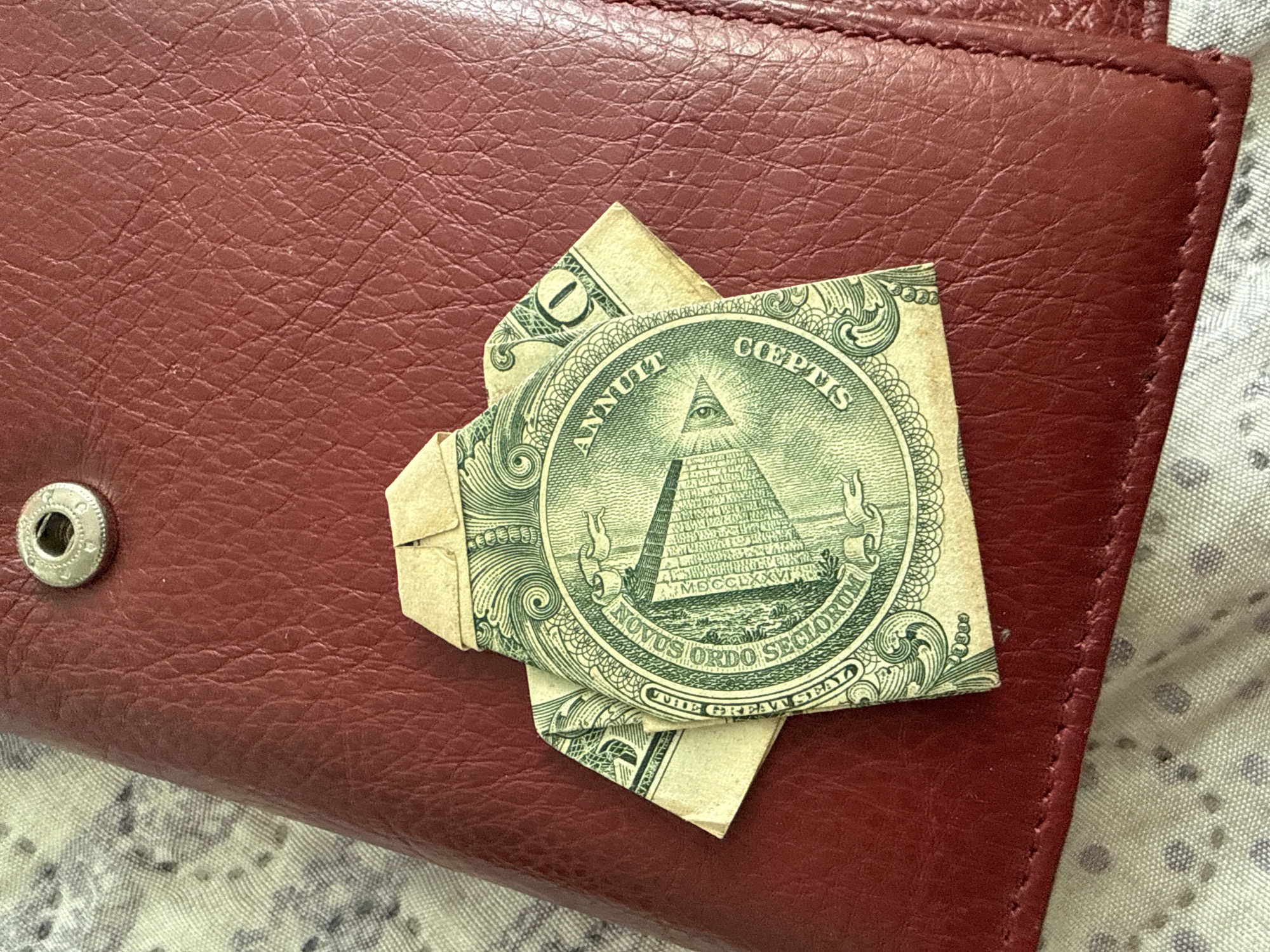 The image displays a United States one-dollar bill folded into the shape of a shirt. The shirt is positioned on a red leather surface, which appears to be part of a wallet or purse. The background shows a textured fabric. The lighting suggests an indoor setting. There are no people or animals visible in the image. There is no information available to place this image in the city of Ekzarh Antimovo, Bulgaria.