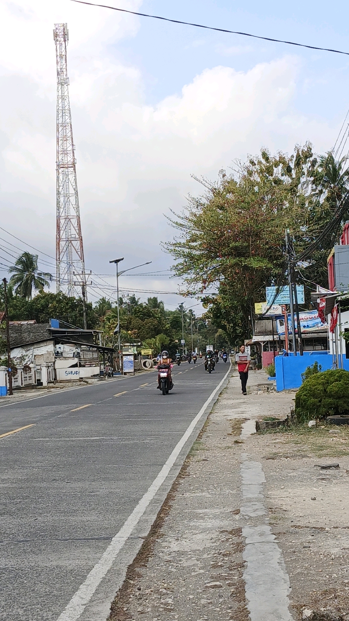 The image depicts a street scene in Cibenda, Indonesia, likely during the day under a cloudy sky. A tall, lattice-style telecommunications tower stands prominently on the left side of the road. The road itself is paved and marked with yellow and white lines. Several motorcycles are visible, with one in the foreground carrying a rider wearing a helmet and a pink top. Further down the road, more motorcycles and riders are present. On the right side of the road, a sidewalk runs alongside the street, with a pedestrian walking away from the camera wearing a red and white striped shirt and dark pants. The environment includes trees with reddish-green foliage and palm trees, as well as some buildings and utility poles with visible wires. There are blue structures and a white building with "SURADY" written on it along the roadside. The ground on the right side of the road is a mixture of gravel and dirt with some concrete elements.