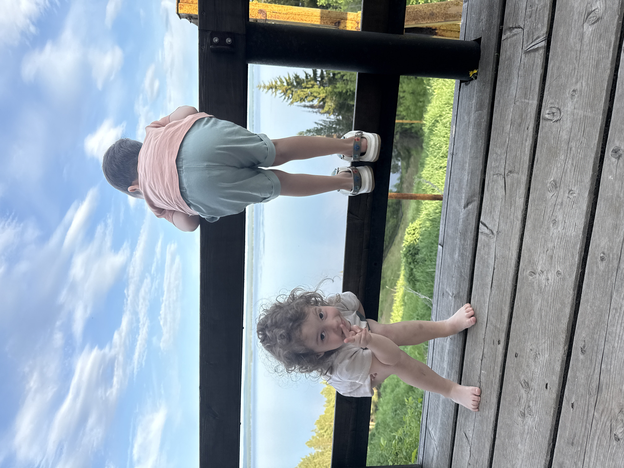 Two young children, one wearing light blue shorts and a pink shirt with white sandals, and the other wearing a white shirt and barefoot, are on a wooden deck looking out over a railing. The scene overlooks water and greenery. The image was taken in Галево, Russia.