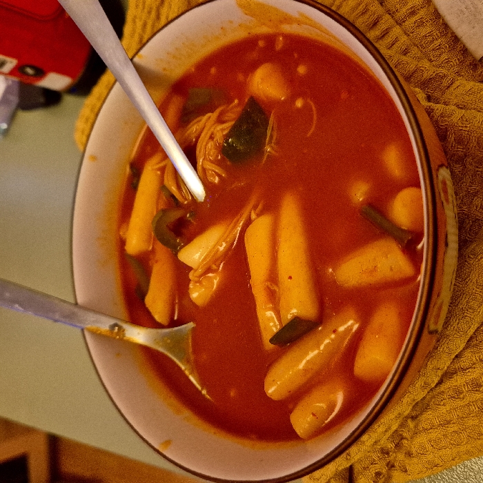 A bowl of Tteokbokki, Korean spicy rice cakes, sits on a yellow waffle-patterned surface. The dish features a vibrant red sauce, with cylindrical rice cakes and green vegetables like scallions and cabbage scattered throughout. Two silver spoons are inside the bowl. A red toy car is partially visible in the upper left. The bowl has a colorful outer pattern with a light-colored interior.