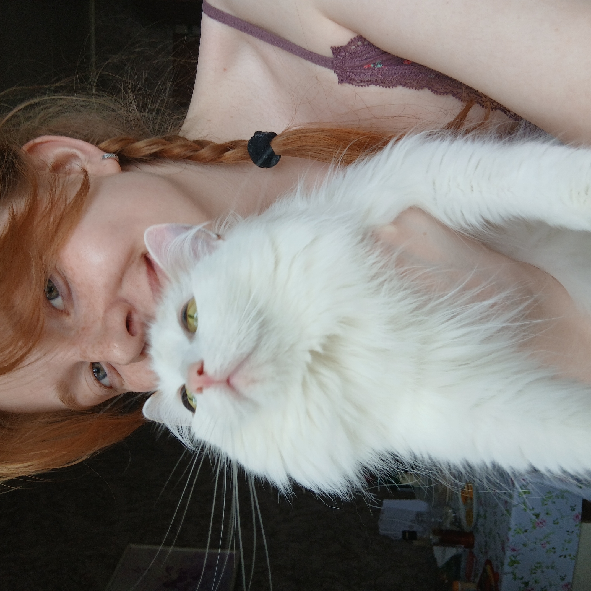 A fair-skinned person with red hair styled in braids is holding a white cat. The person is wearing a brown bra. The cat has green eyes. The background includes a dark brown surface and a table with a floral tablecloth. The location is not discernible from the image.