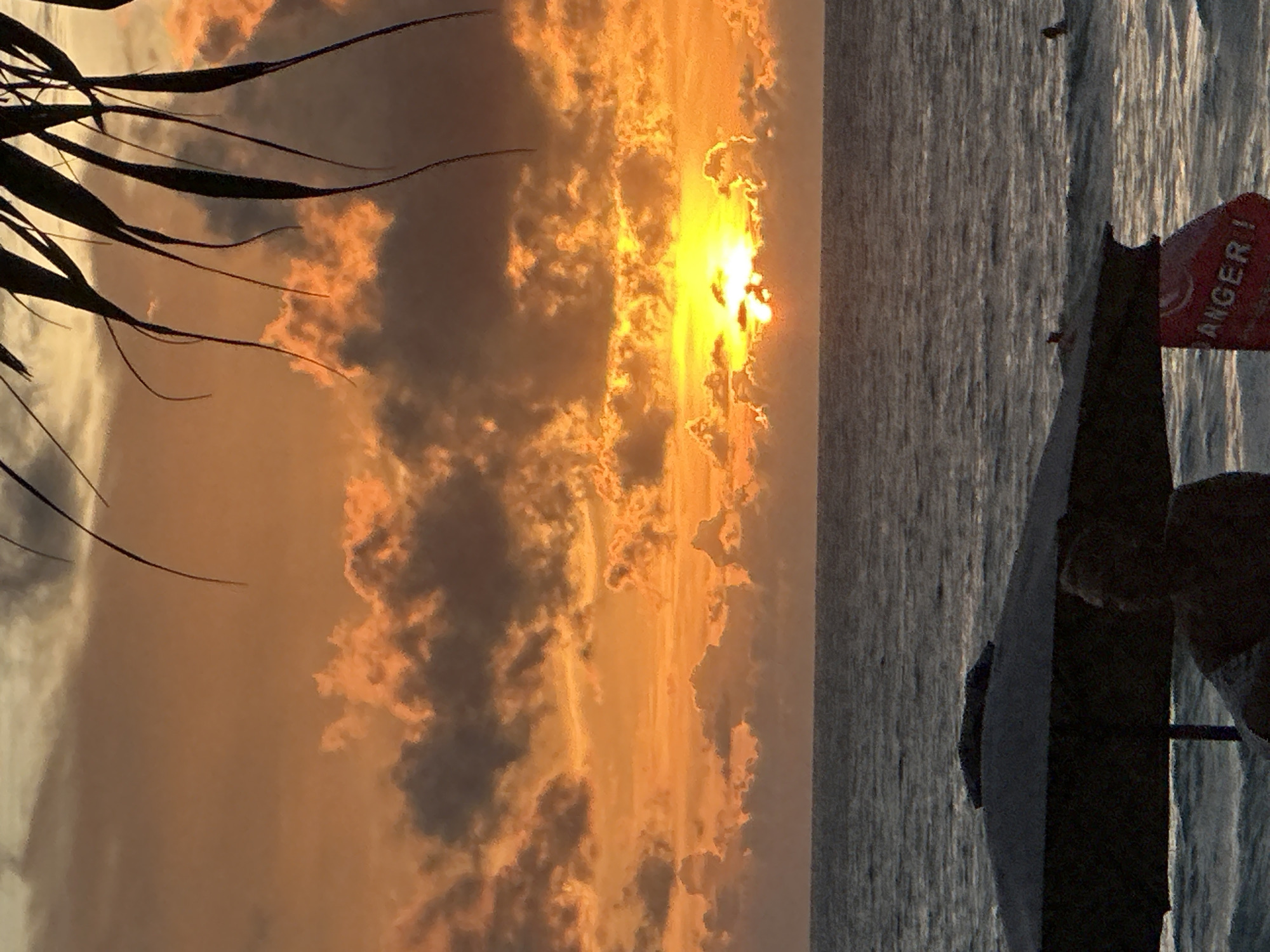 A wide shot captures a sunset over the ocean in Karon, Thailand. The sky is filled with hues of orange and yellow, with dramatic clouds lit by the setting sun. Palm fronds hang from the top of the frame, silhouetted against the vibrant sky. In the foreground, two beach umbrellas are visible, one dark and the other with a red "ANGER" sign. A person is partially visible sitting near the umbrellas. The ocean stretches out to the horizon, with gentle waves and a few distant swimmers.