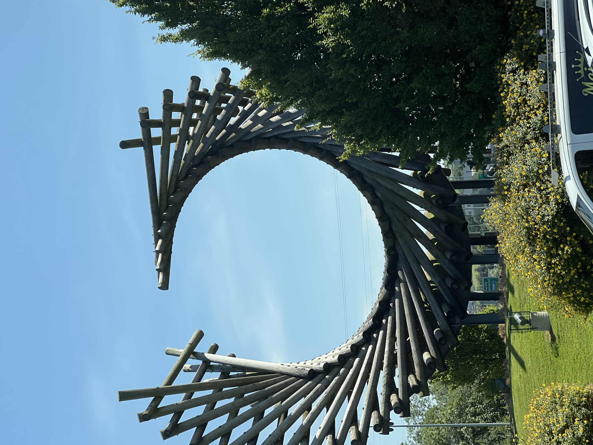 The image shows a large, circular wooden sculpture in Letterkenny, Ireland, against a clear blue sky. The sculpture is made of numerous wooden poles arranged in a spiral pattern, creating an open, circular form with a gap in the middle. The sculpture is situated on a grassy mound, with a dense green tree on the right side of the frame partially obscuring the sculpture's right edge. In the foreground, there are bright yellow flowering bushes and a white van is partially visible at the bottom. A signpost is visible in the background, indicating "Troy Edge" and "HER Routes." The sculpture appears to be a public art installation.