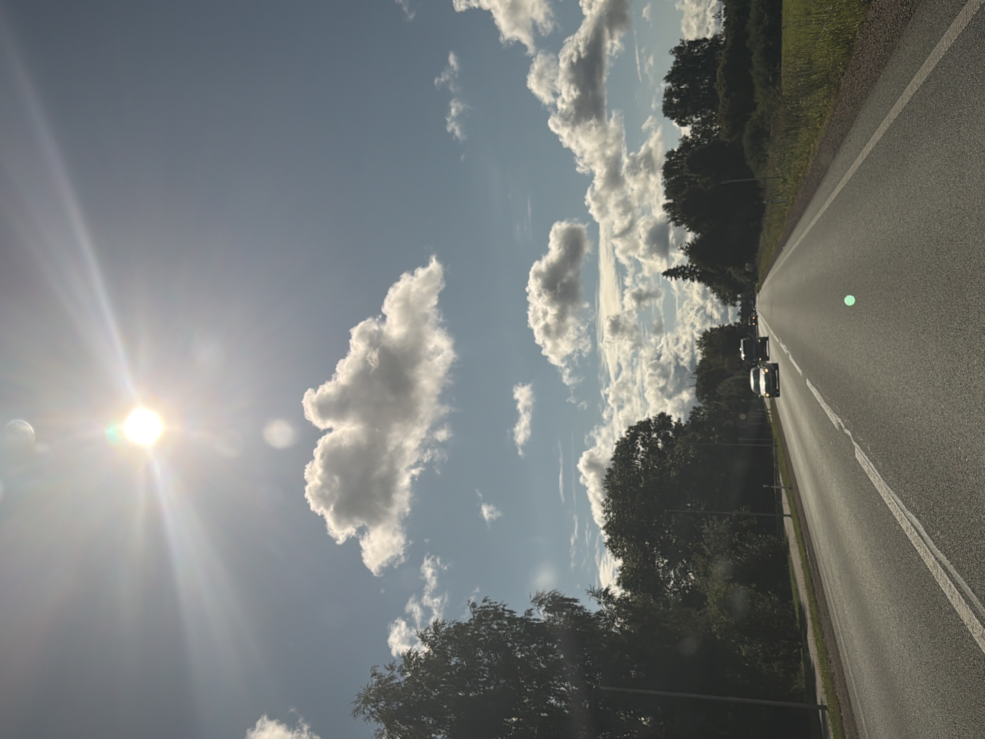 A forward-facing, wide-angle, eye-level view shows a two-lane asphalt road in Latvia, likely in or near Ķekava, on a sunny day. The sun is bright and high in the sky, creating lens flare and a bright halo. There are scattered white cumulus clouds against a light blue sky. Tall, dark green trees line the left side of the road, with a field of green grass and more trees visible on the right. A few cars are traveling on the road in the same direction as the viewer. The road has solid white lines marking the lanes and a dashed white line on the right edge. The overall scene suggests a rural or suburban environment with natural surroundings and moderate traffic.