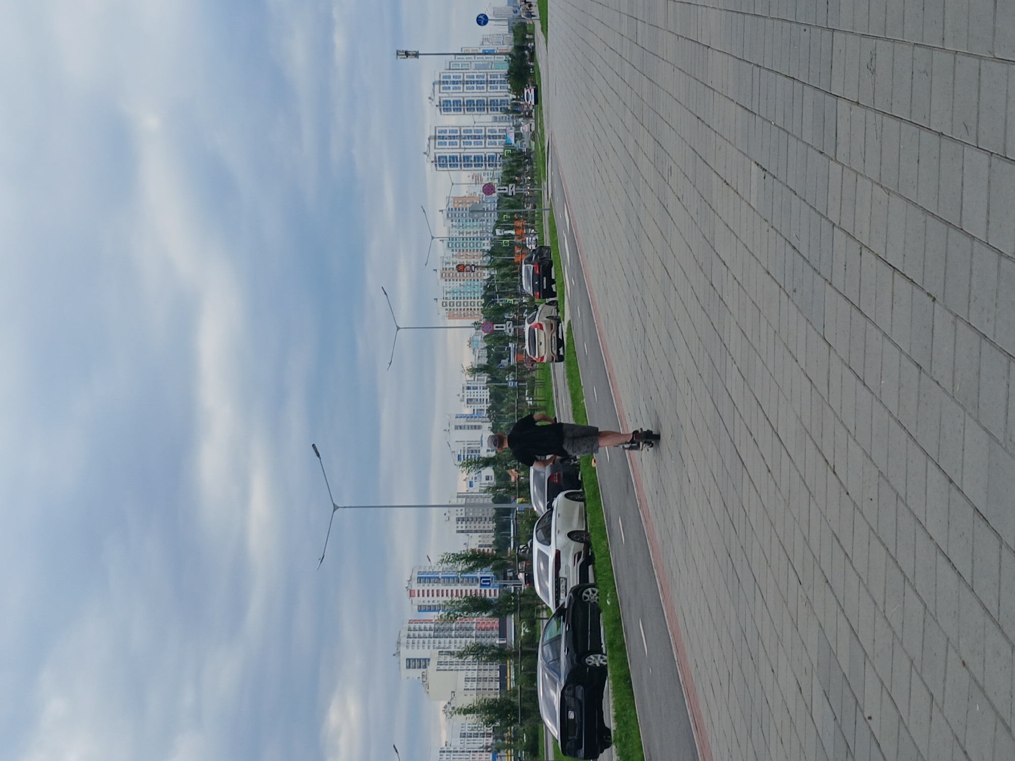 In Yekaterinburg, Russia, a man rides an electric scooter on a paved pedestrian path adjacent to a road with parked cars and a bike lane. The man is wearing a black t-shirt and shorts, and a grey cap. In the background, a row of modern, multi-story apartment buildings with diverse architectural styles stands under a cloudy sky. Streetlights are visible along the road.