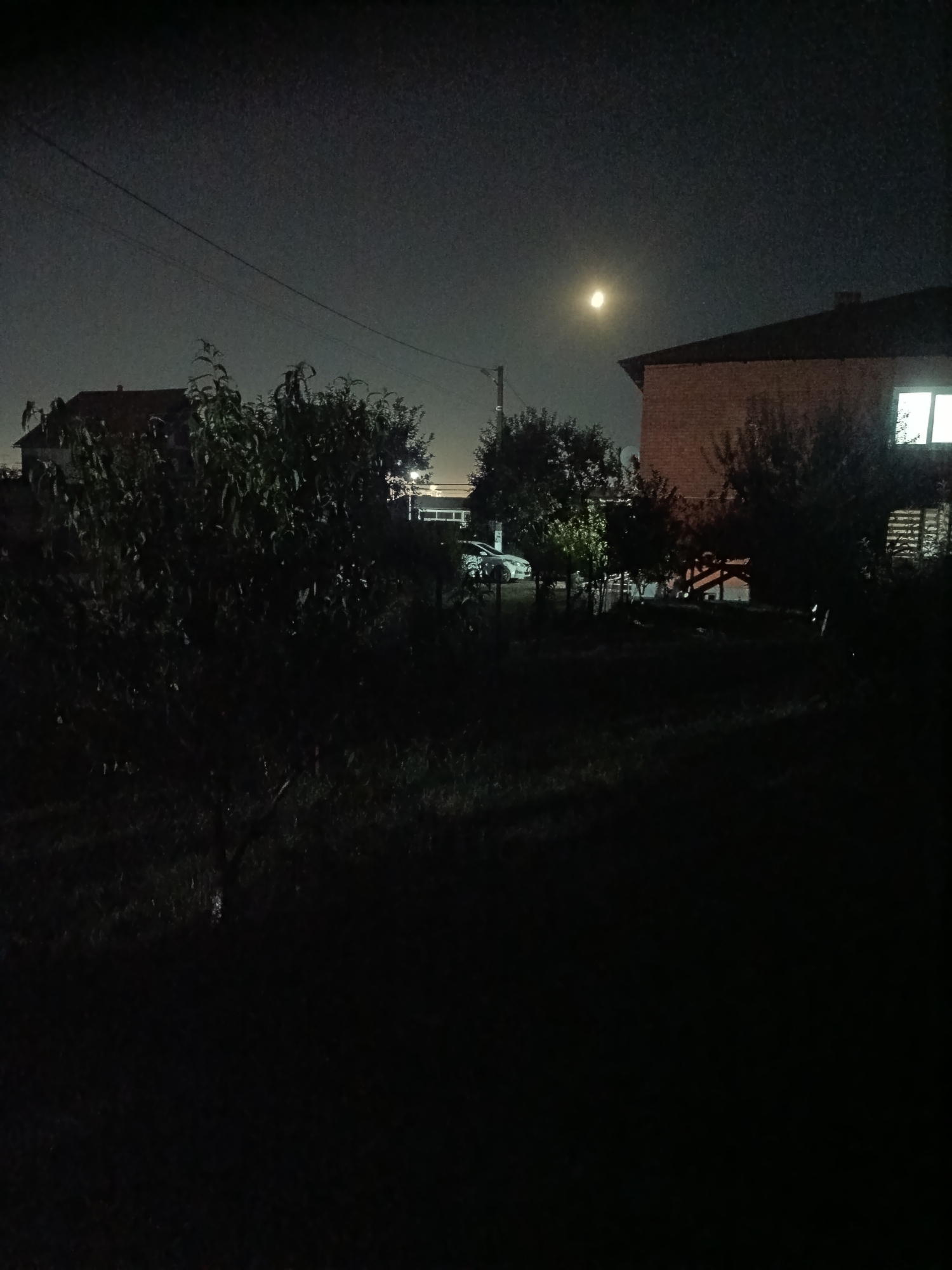 A nighttime, wide shot captures a residential scene with a pale moon high in the dark sky. The foreground is dominated by deep shadows, obscuring much of the ground and vegetation. Two houses are visible. On the left, a darker, brick building with a pitched roof is partially illuminated by ambient light. On the right, a lighter-colored brick building features a prominent lit window. A car, possibly white, is parked in the middle ground, partially obscured by trees and shadows. Trees and bushes are scattered throughout the scene, their forms indistinct in the low light. Power lines stretch across the upper portion of the image. The overall impression is one of a quiet, suburban night.
