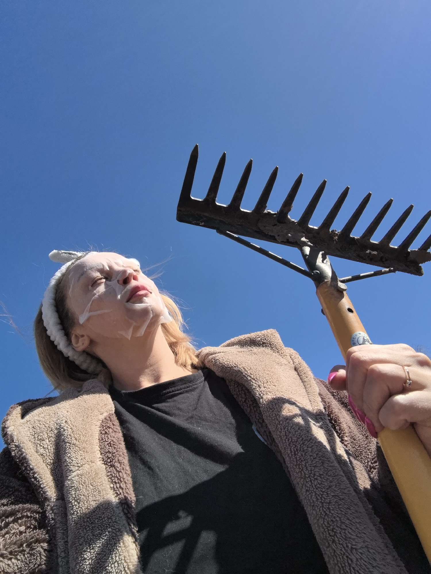 A woman with a white facial mask and a headband with bunny ears is holding a rake in her right hand. She is wearing a black t-shirt and a brown and beige hooded jacket. The background is a clear blue sky. The location is not discernible from the image.