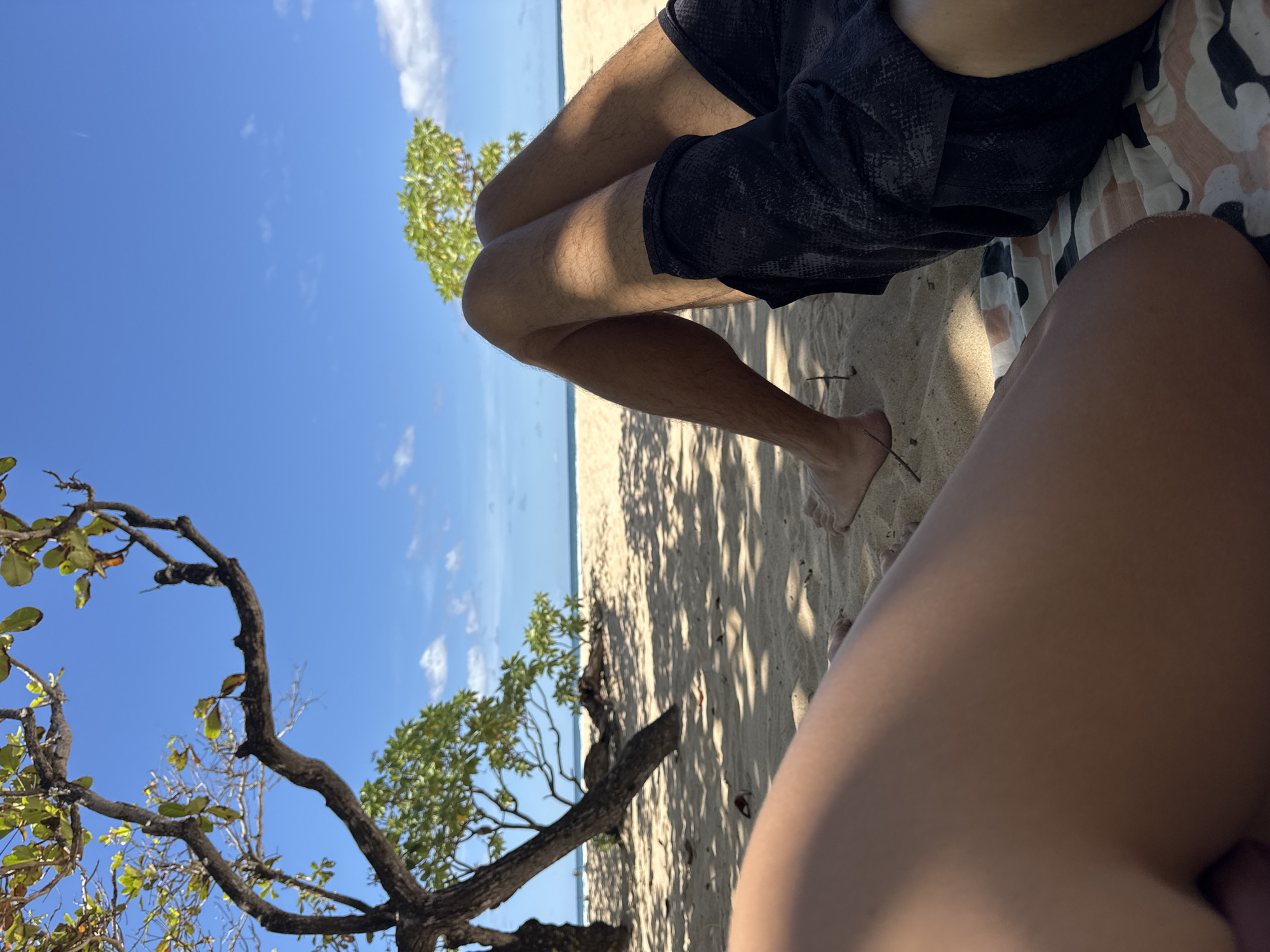 The image captures a beach scene in Kuta Selatan, Indonesia, from a first-person perspective. The observer is seated on a sandy beach, likely on a patterned cloth. A person is lying next to the observer, wearing dark shorts. The observer's legs are visible in the bottom right corner of the image. In the background, there is a sandy beach and the ocean with a visible horizon line. Trees with green foliage are scattered across the beach. The sky is blue with scattered clouds. The shadows of trees are cast on the sand.