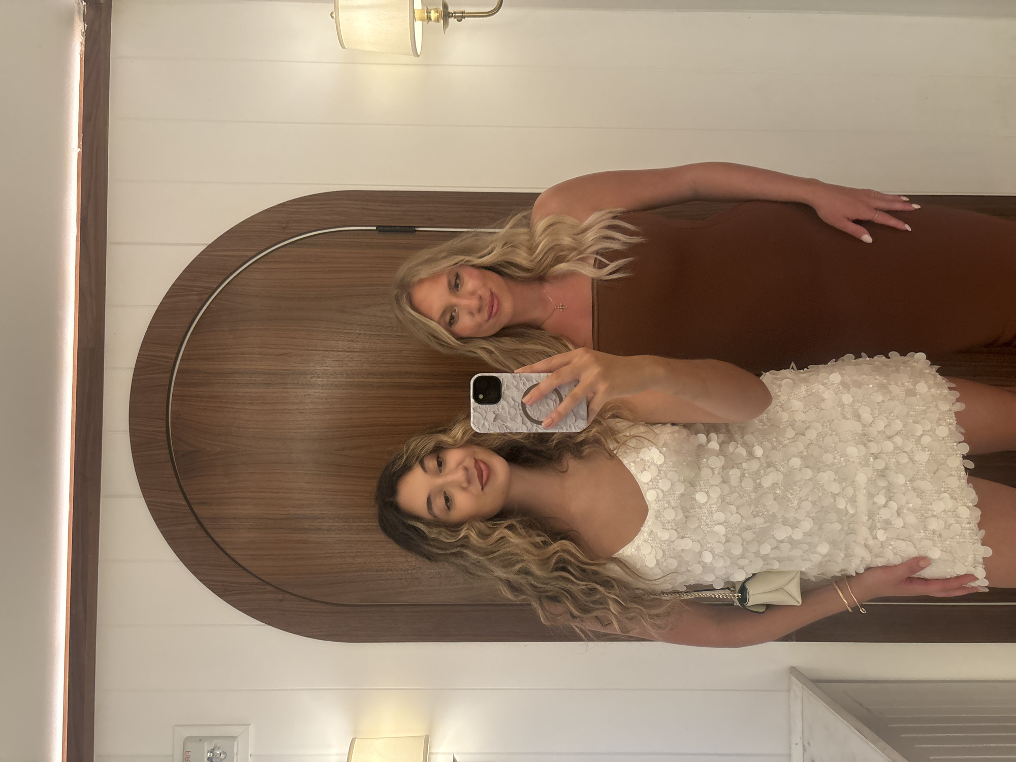 Two women are taking a mirror selfie in a bathroom. The woman on the left is wearing a white sequined mini dress and the woman on the right is wearing a brown strapless midi dress. The background features a large wooden arched doorway and white paneled walls. A light fixture is visible on the right wall. The scene appears to be a hotel bathroom in Miami, Florida.