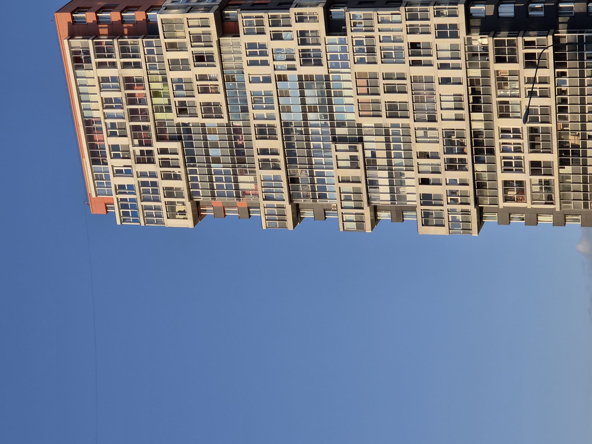 A close-up, vertical shot focuses on the right side of a modern, multi-story apartment building against a clear, bright blue sky. The building's facade is composed of numerous rectangular windows, many with white frames, interspersed with sections of light beige or grey material. Some windows reveal glimpses of indoor curtains, and a few are open. The building's design features varied projections and recesses, creating an asymmetrical and dynamic appearance. The upper section of the building is accented with a reddish-orange color. A street lamp with a curved arm is visible in the lower right quadrant, its pole extending upwards. A thin, dark line, possibly a wire, is faintly visible horizontally across the sky in the upper left portion of the image. The setting appears to be an urban environment in Kudrovo, Russia. There are no visible people or specific actions captured in this static scene.
