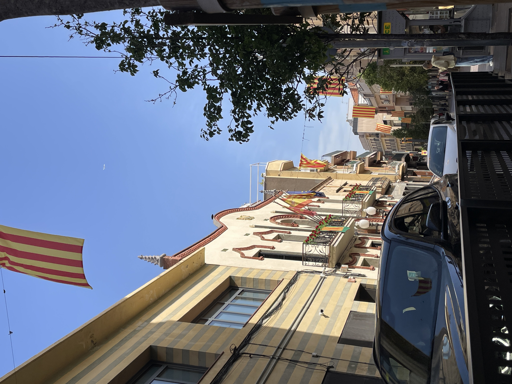 A street in Montcada, Spain is lined with buildings adorned with Catalan flags, indicating a possible cultural event or celebration. A tree with dense foliage hangs overhead. Two vehicles, a blue car and a white van, are parked on the right side of the street. People are walking along the sidewalk in the distance. The architecture of the buildings is distinct, with striped patterns and decorative elements.