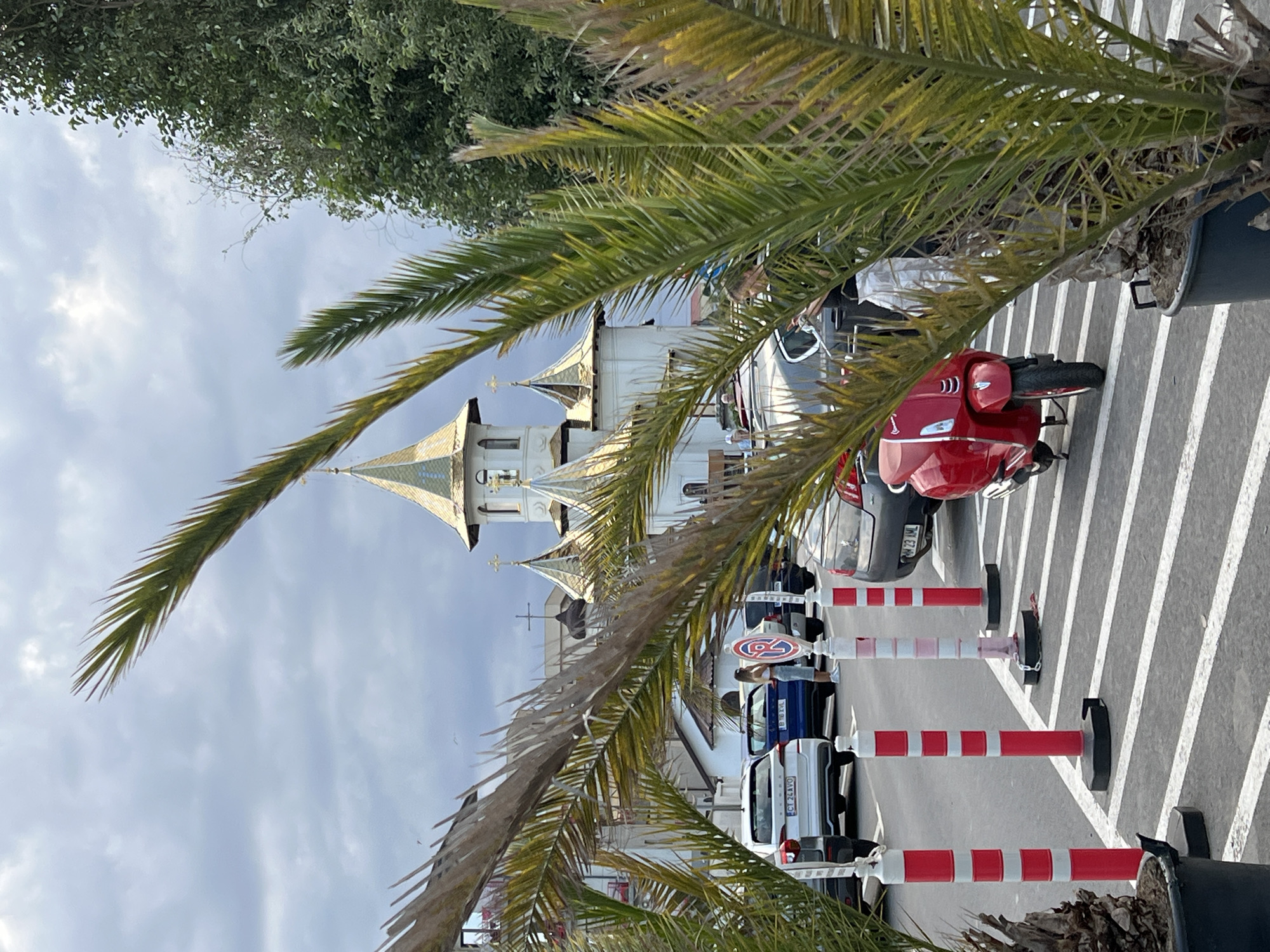 In Năvodari, Romania, this image captures a street scene with a prominent church building featuring golden domes. In the foreground, palm trees frame the view, with one on the right obscuring parts of the frame. A red scooter is parked on the right side of the street, positioned on white painted parking lines. Several cars are parked in the background, including a white Skoda and a dark blue vehicle. A woman in a white top and skirt is standing near the blue car, facing away from the camera. Another woman in a black top is visible behind the palm tree on the right, looking at her phone. Red and white bollards are placed along the parking spaces. The sky is overcast with clouds.
