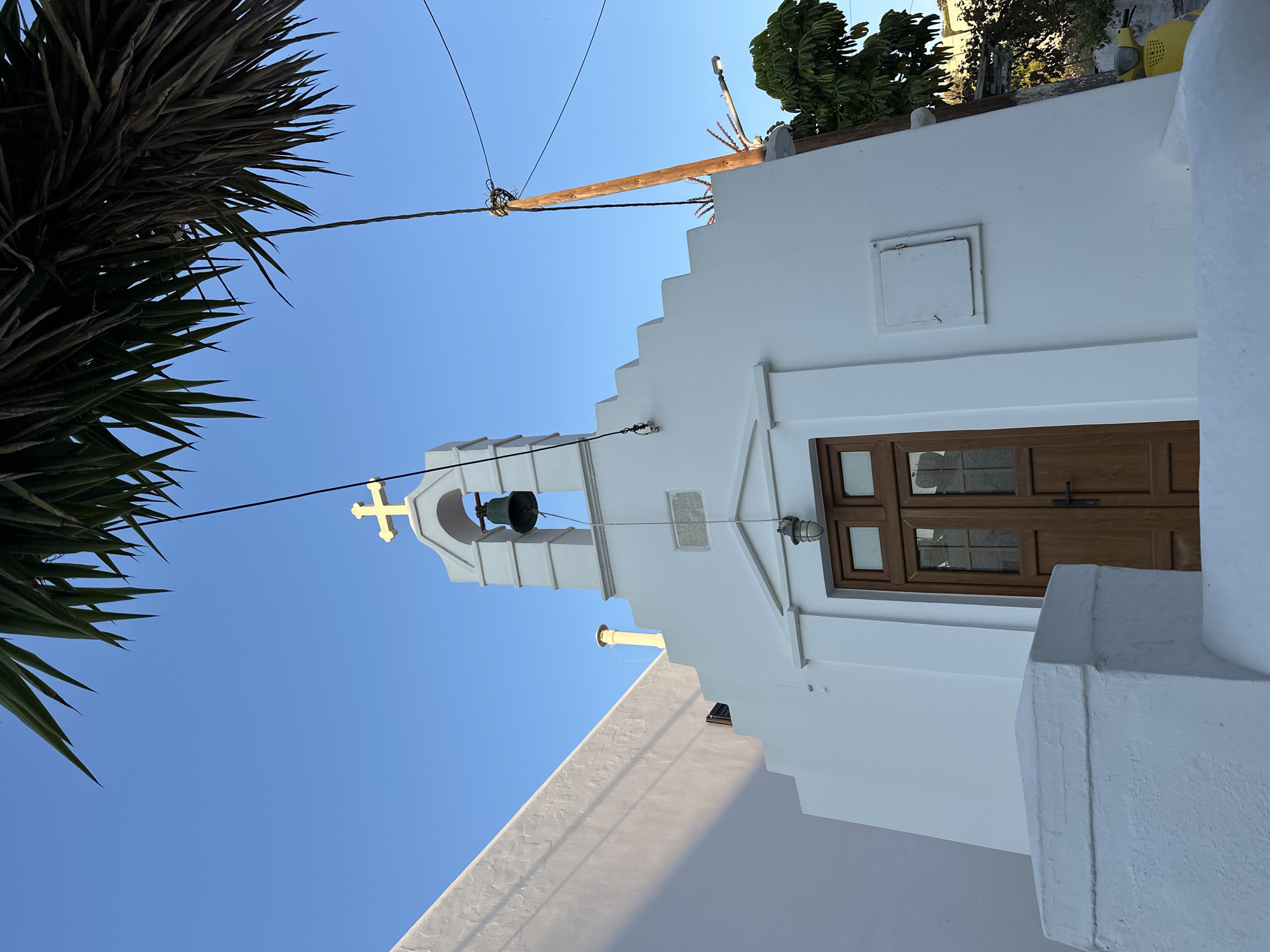 In Mykonos, Greece, a white church with a bell tower and a golden cross is pictured under a clear blue sky. Palm fronds hang over the scene from the top of the frame. A wooden door with glass panels is centrally located on the church's facade, below a small plaque. Electrical wires and a wooden utility pole are visible to the right of the church, along with a yellow scooter parked in the background. The architecture features stepped rooflines and a white railing in the foreground.