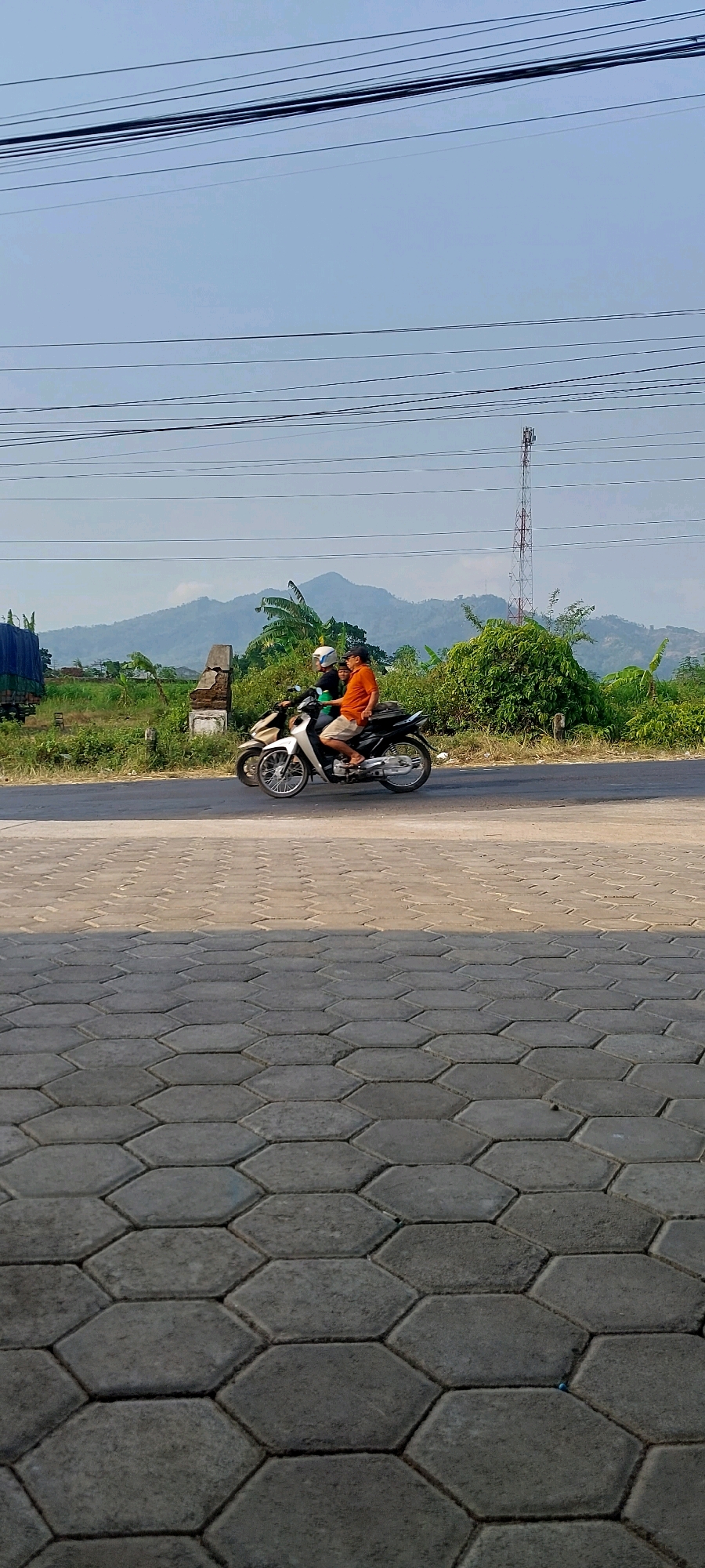 The image captures a sunny outdoor scene on a paved road in Keling, Indonesia. Two individuals are riding a motorcycle, with the person in the front wearing a white helmet and the passenger in the back wearing an orange shirt and a dark cap. The motorcycle is light-colored with black accents and has a visible silver exhaust pipe. They are traveling on a paved road that runs alongside a grassy area with lush green foliage and distant mountains. Several power lines are visible crisscrossing the sky, and a cell tower stands tall in the background. The foreground of the image is dominated by a hexagonal-patterned interlocking concrete paver surface, casting a distinct shadow. The time of day appears to be late morning or early afternoon, given the sunlight and shadows.