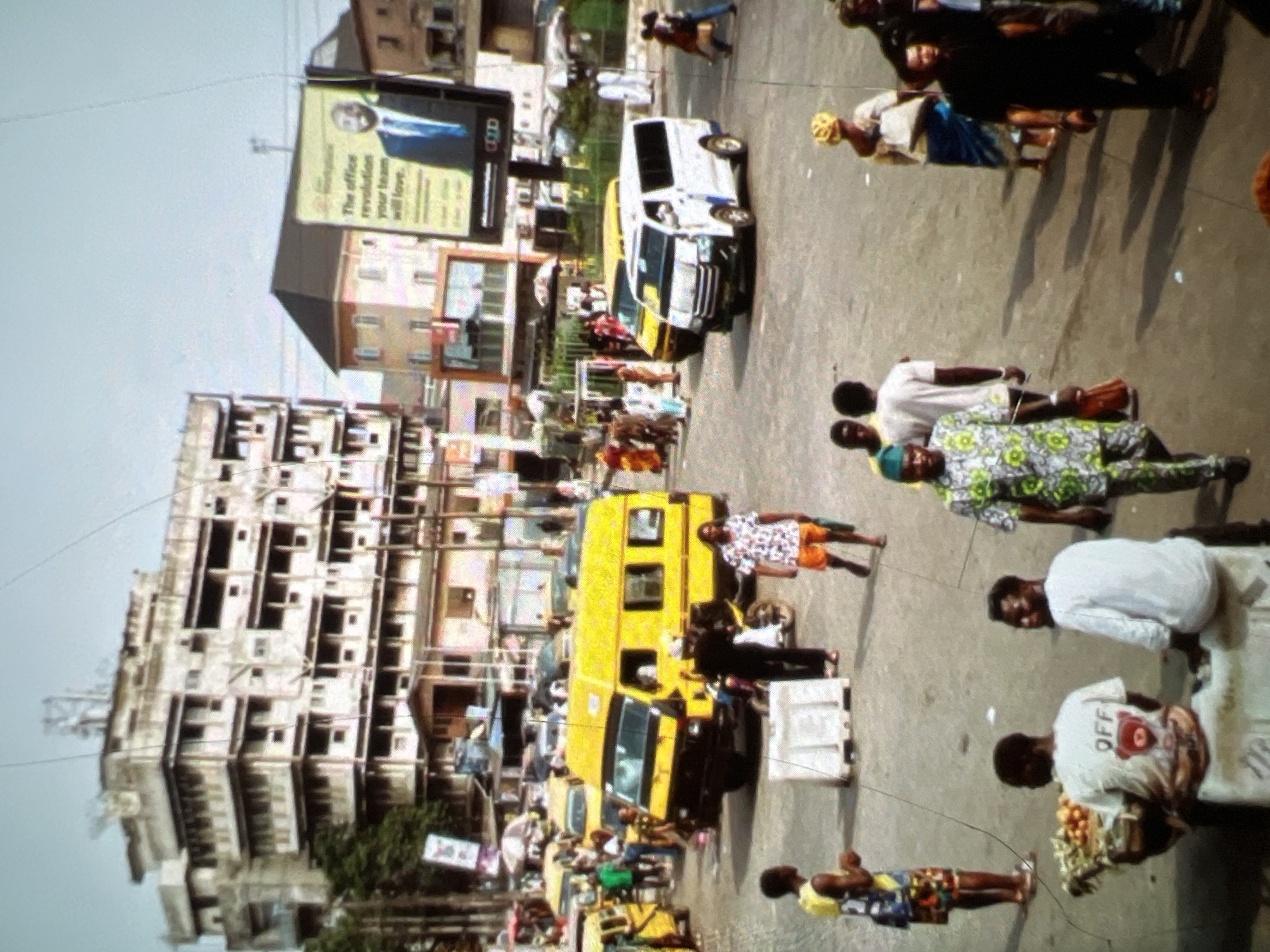 The image depicts a street scene in Lagos, Nigeria, characterized by a busy intersection with several vehicles, pedestrians, and buildings. A prominent yellow minibus, commonly known as a danfo, is visible in the foreground, suggesting public transportation is in operation. Adjacent to it, a white minibus also appears to be a form of transport. The street is paved, and there are pedestrians walking in various directions, some carrying items. Buildings line the background, some appearing to be older structures, while a billboard with an advertisement is also present. The overall atmosphere suggests a bustling urban environment.